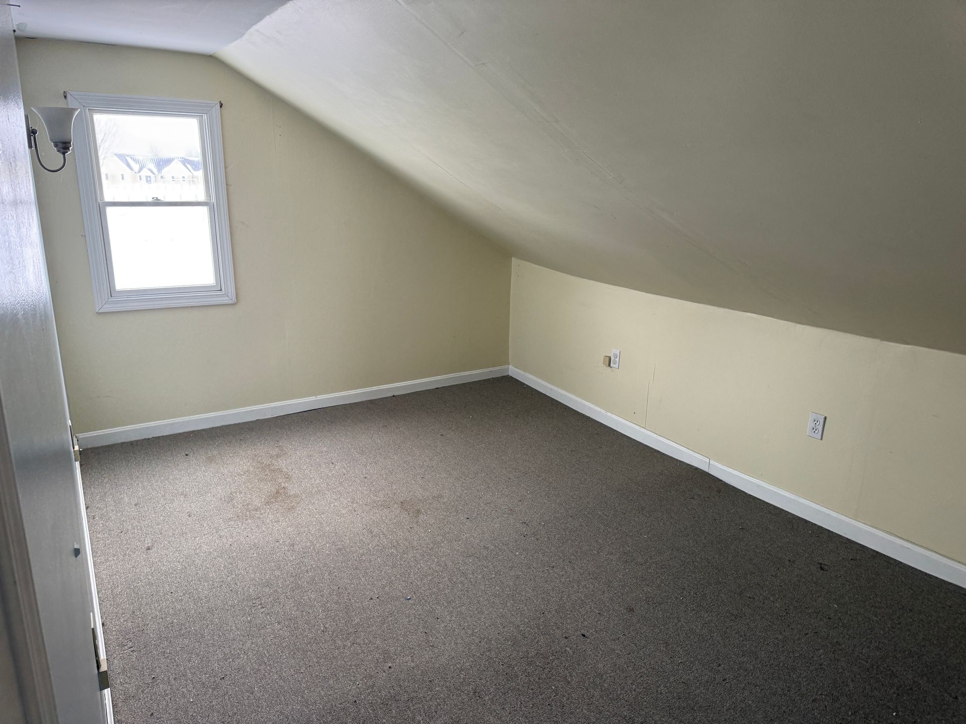 Empty attic room with tan walls, slanted ceiling, small window, and brown carpet.