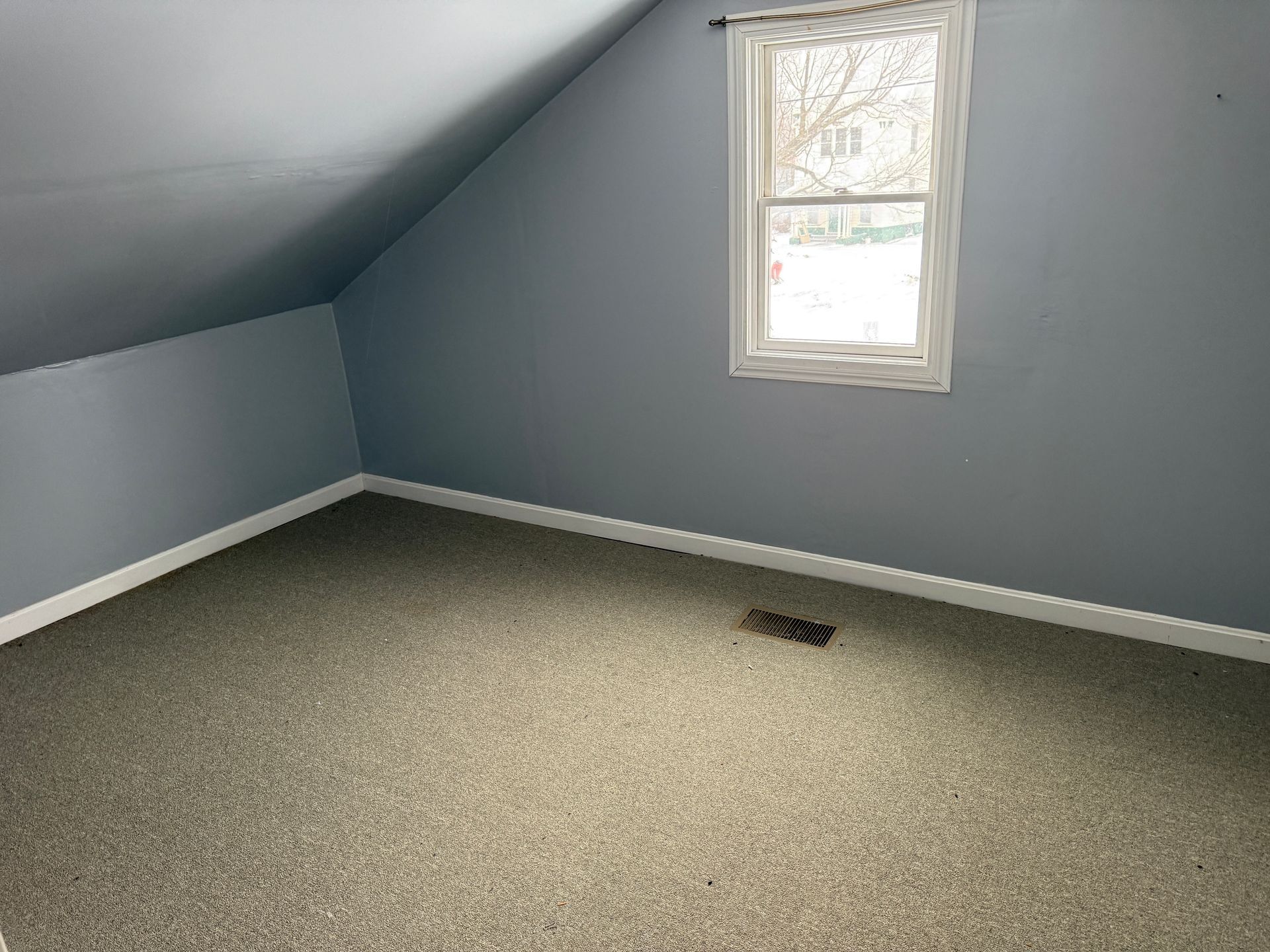 Empty room with gray walls, angled ceiling, small window, and speckled carpet.