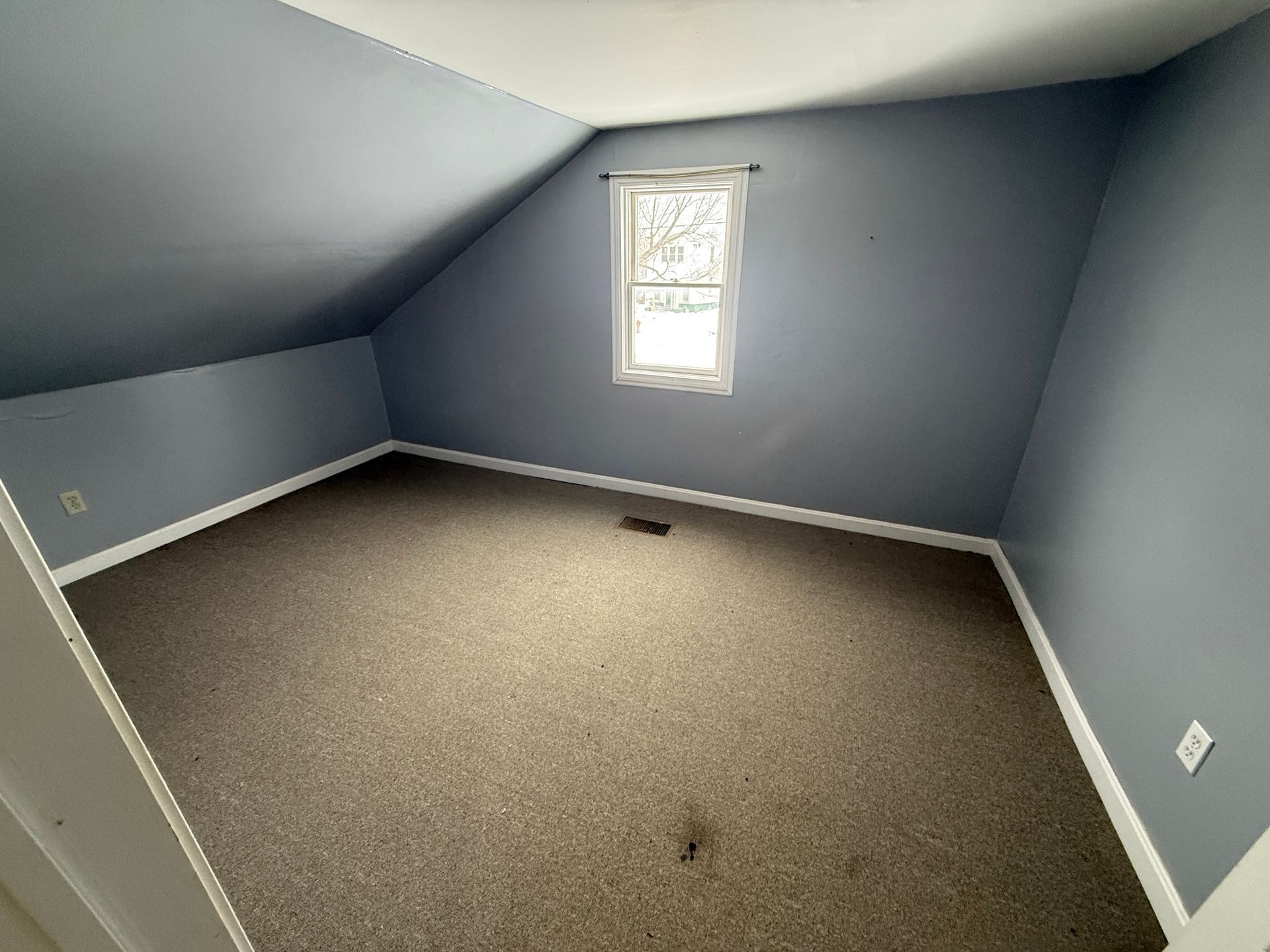 Empty room with angled ceilings, small window, and brown carpet; walls are blue.