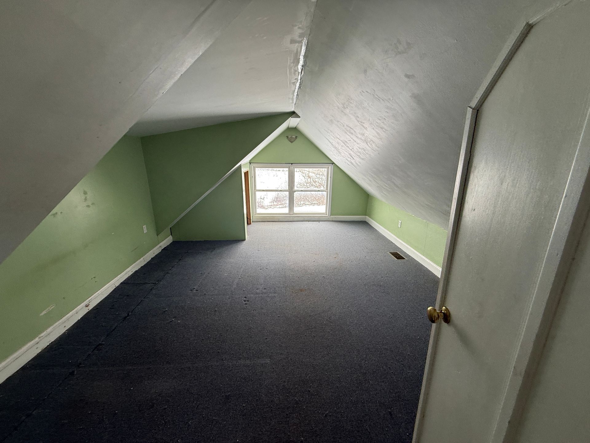 Attic room with sloped ceiling, blue carpet, green walls, and a small window. White door on the right.