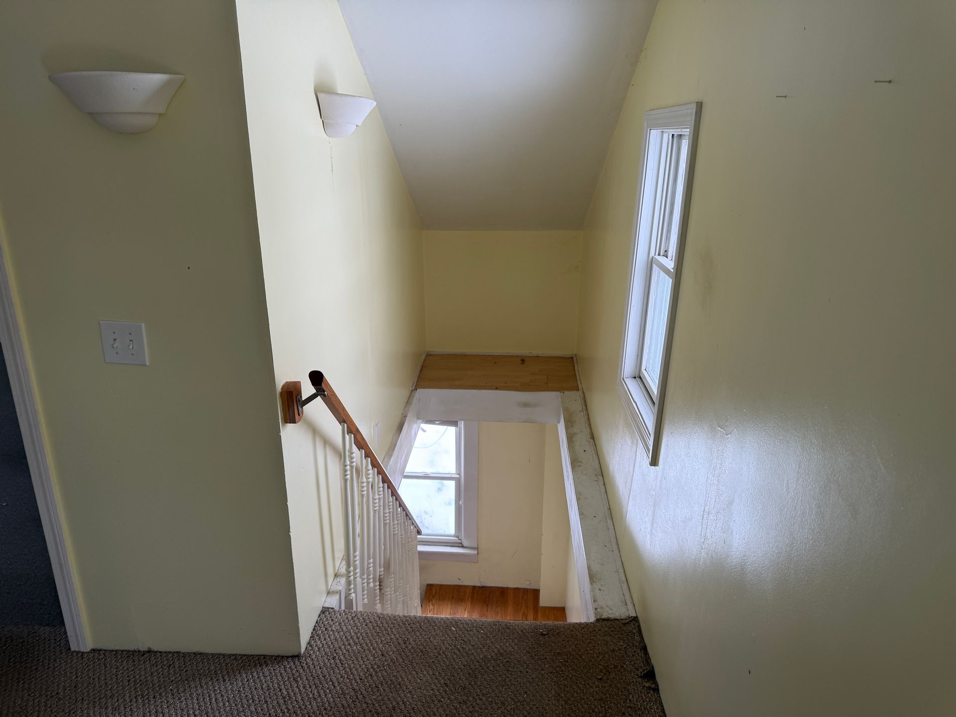 Staircase with light-colored walls and a window at the bottom. The stairs have a brown carpet and white railing.