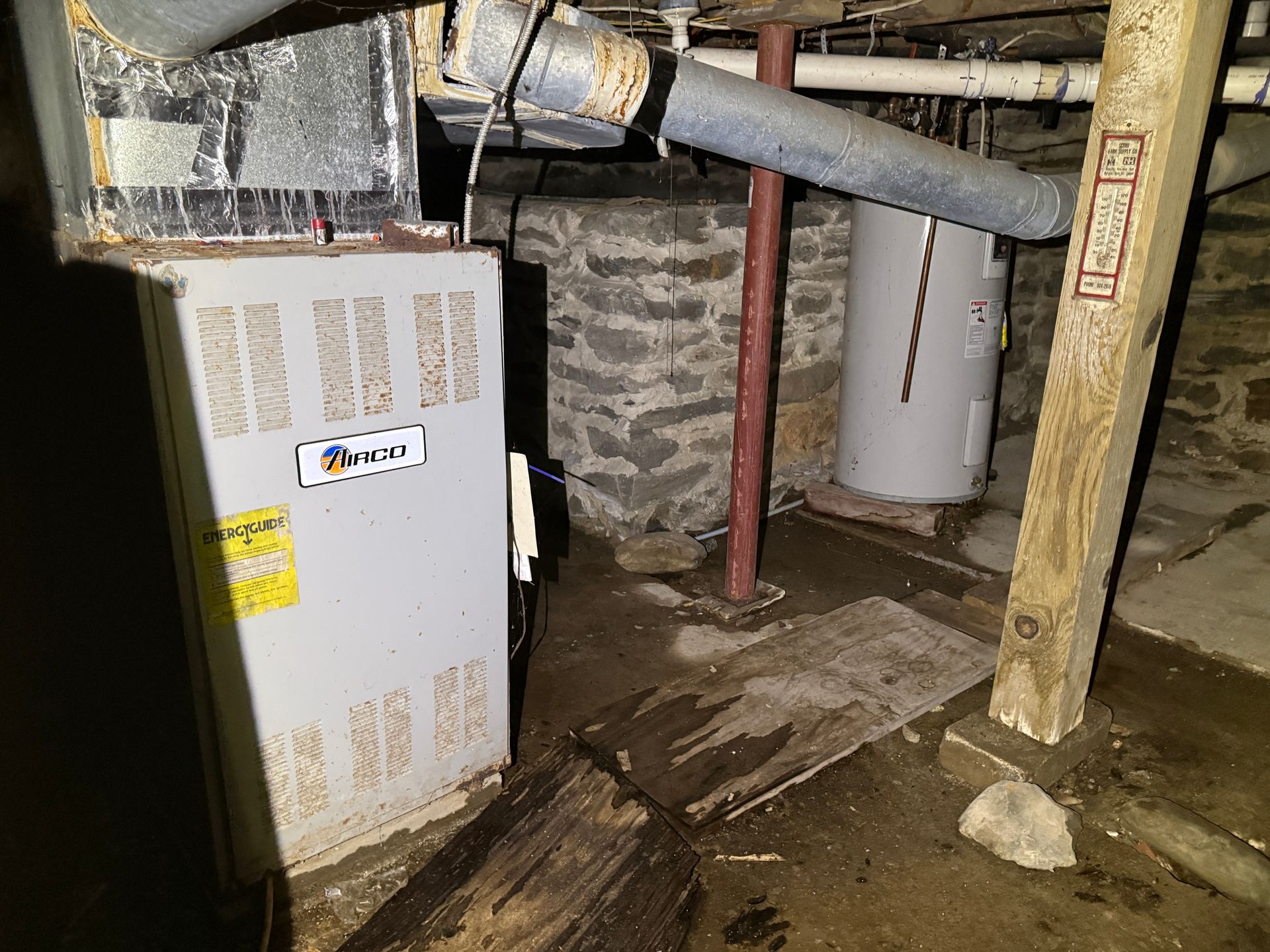 Furnace and water heater in a basement with stone walls and wooden support beams.