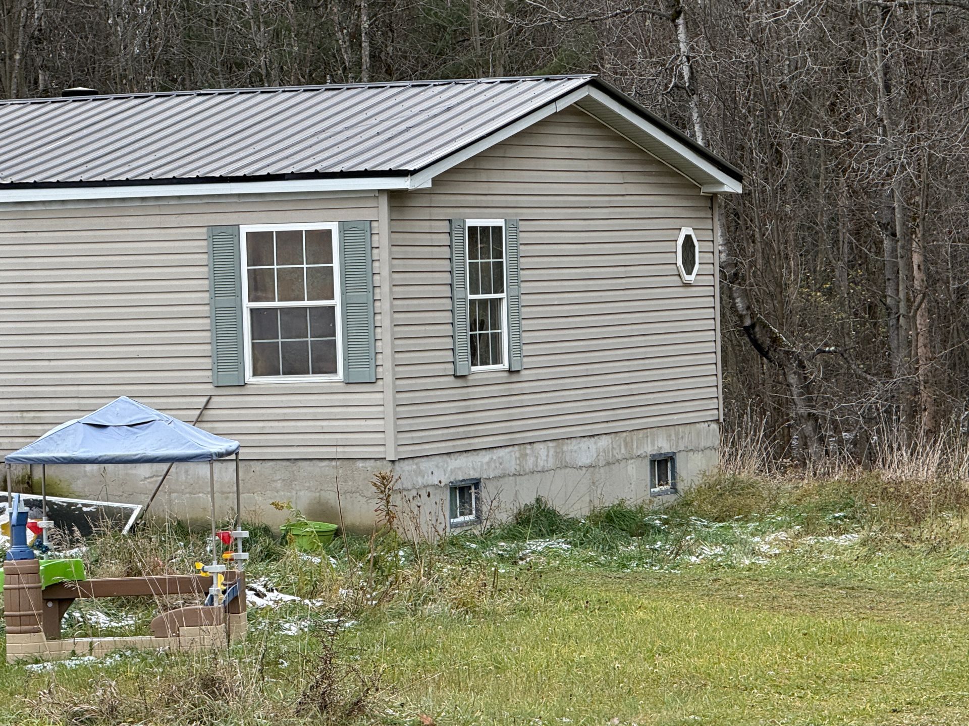 Small beige house with a gray roof beside a wooded area