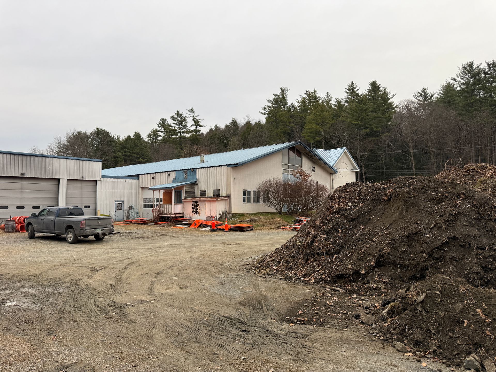 Building with blue roof, work vehicles, and large dirt pile. Exterior shot, overcast day.