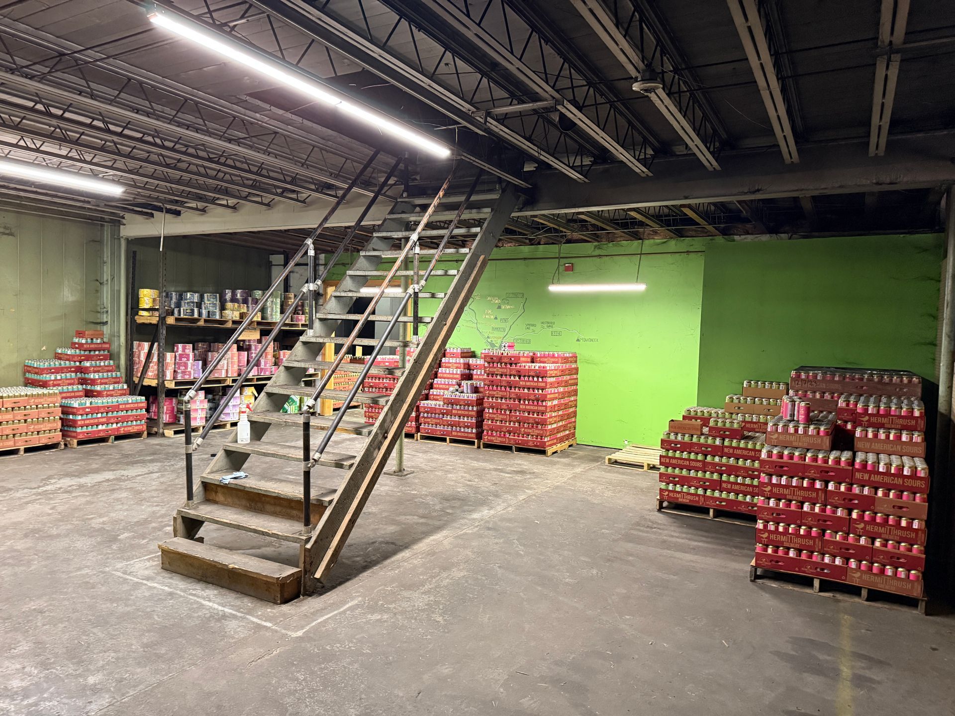 Warehouse interior with stacked goods, metal stairs, and overhead lighting.