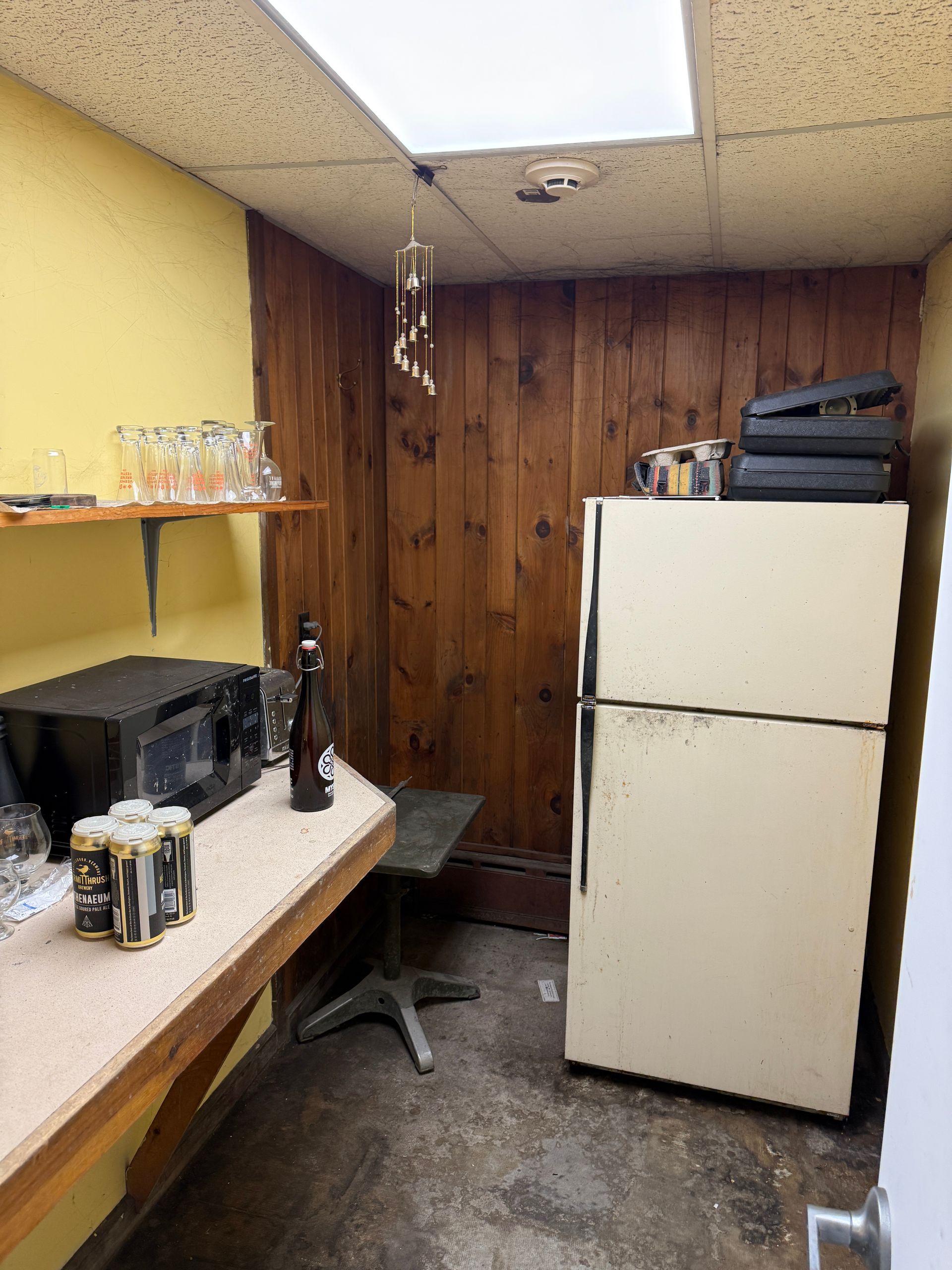 Small kitchen area with fridge, microwave, shelving, and a stool. Yellow and wood-paneled walls, concrete floor.