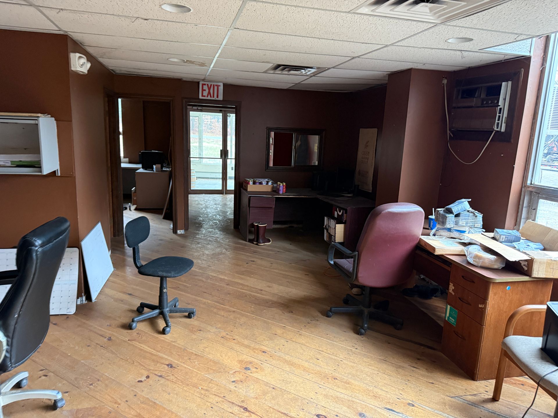 Interior of a brown-walled office, empty desks, chairs, and materials around. Exit sign visible.