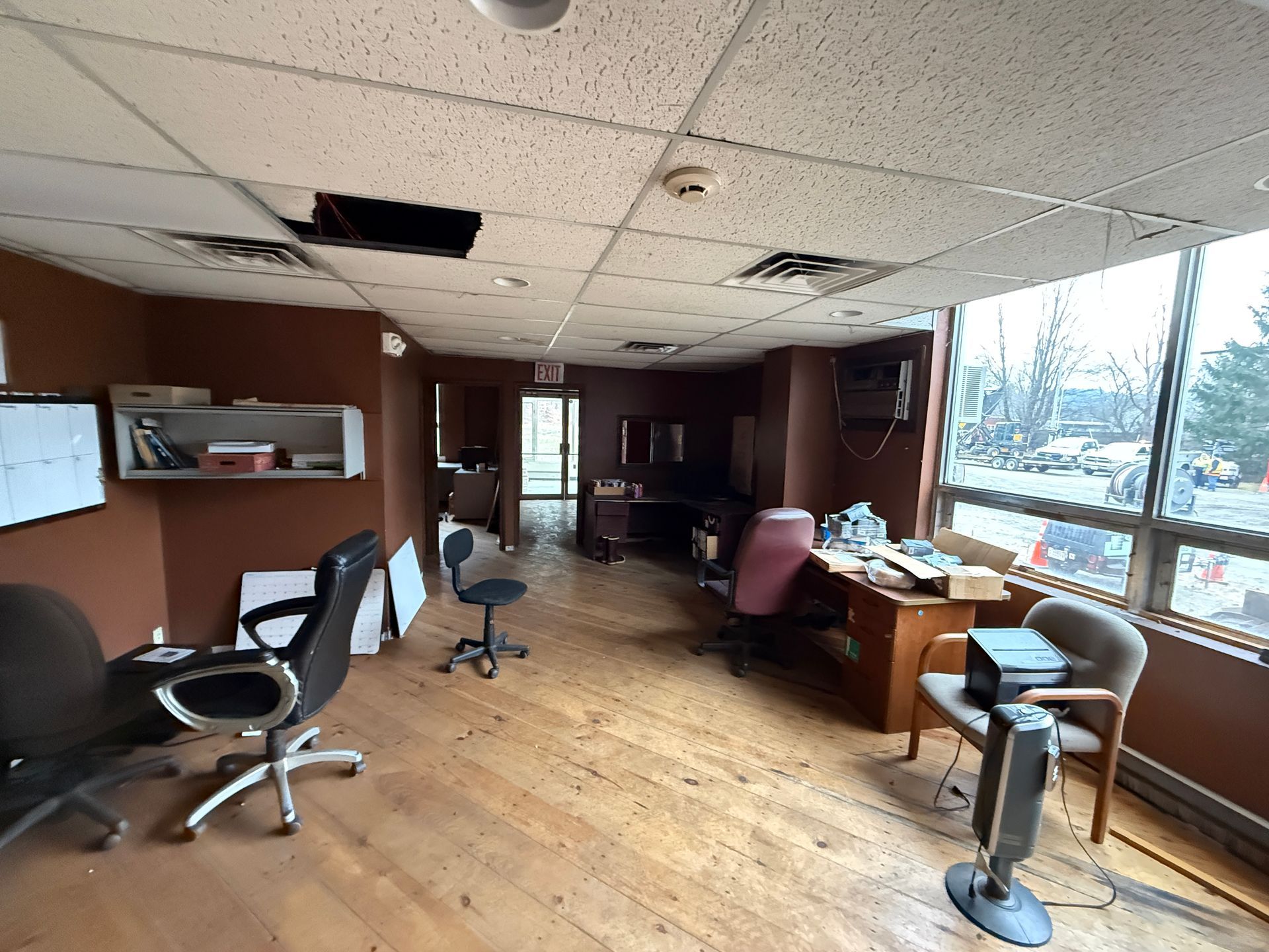 Office interior with ceiling, brown walls, and wooden floor. Desks, chairs, and a window.