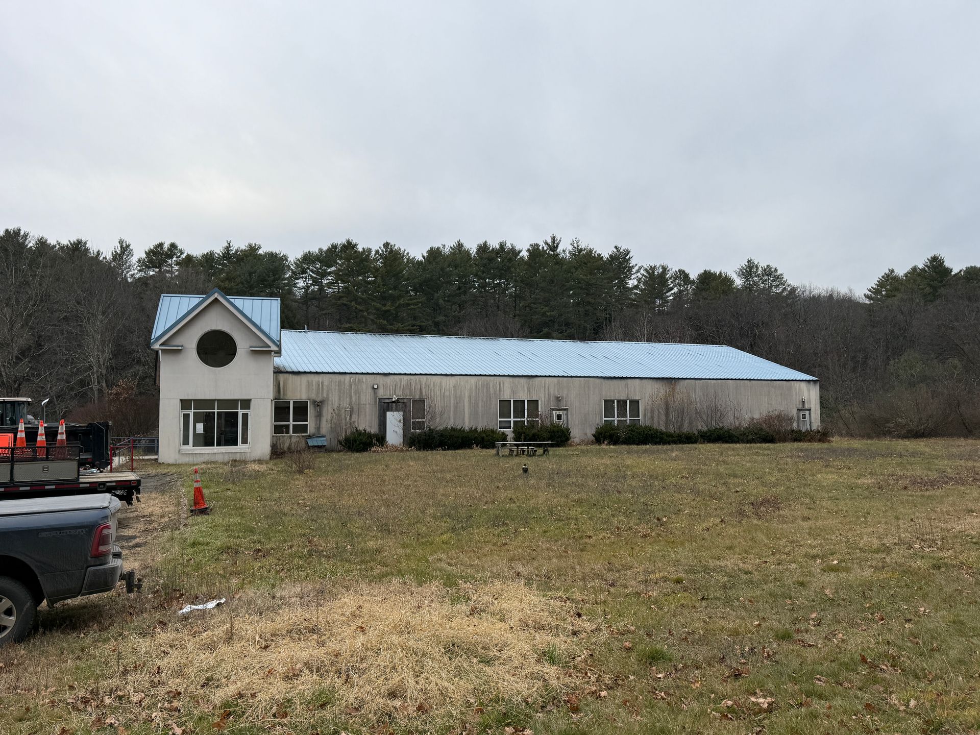 Building with a blue roof and round window on the left, set in a grassy field, trees in the background, cloudy sky.