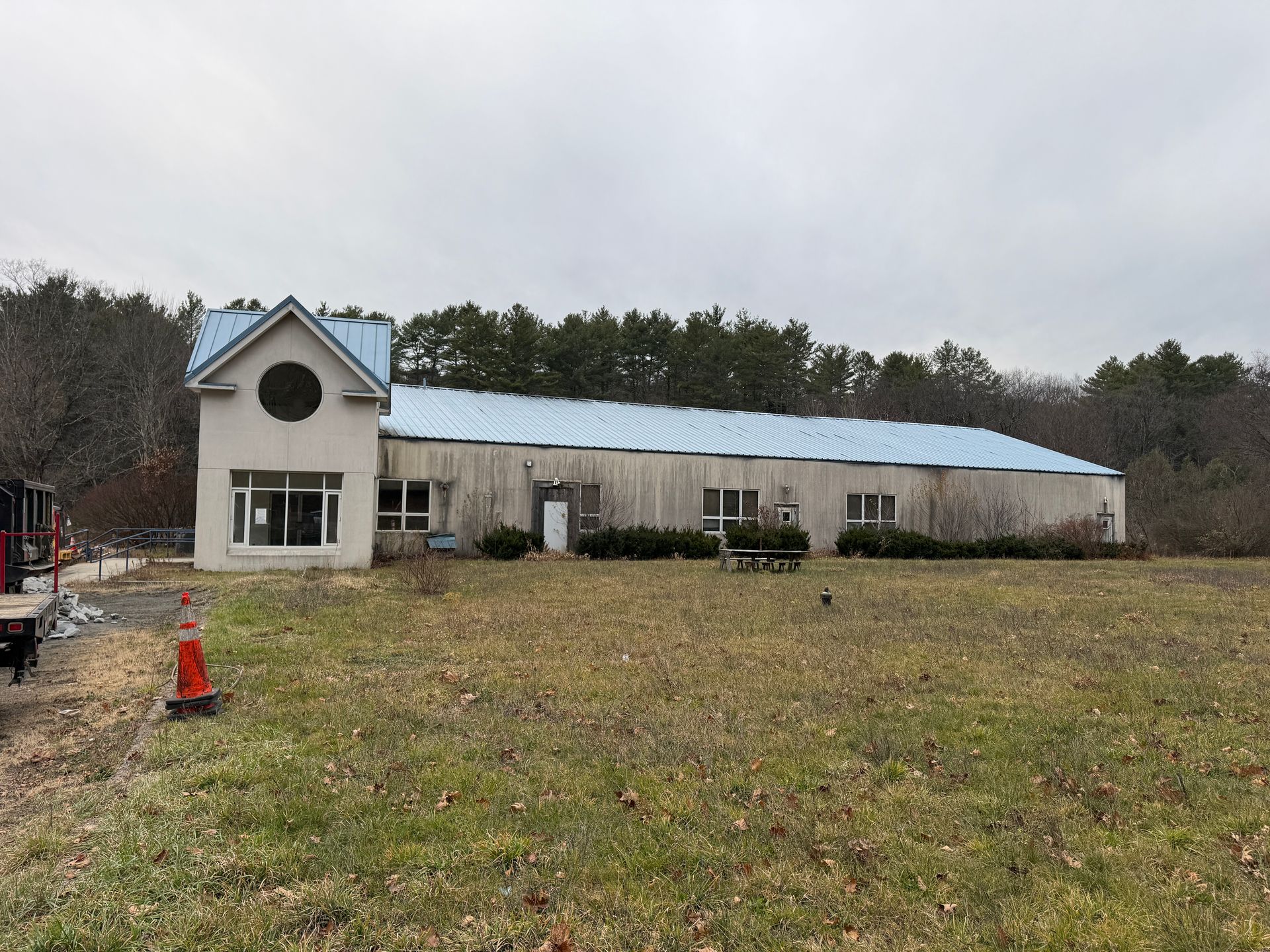 Warehouse building with blue roof and circular window, on a grassy field with trees in the background under an overcast sky.