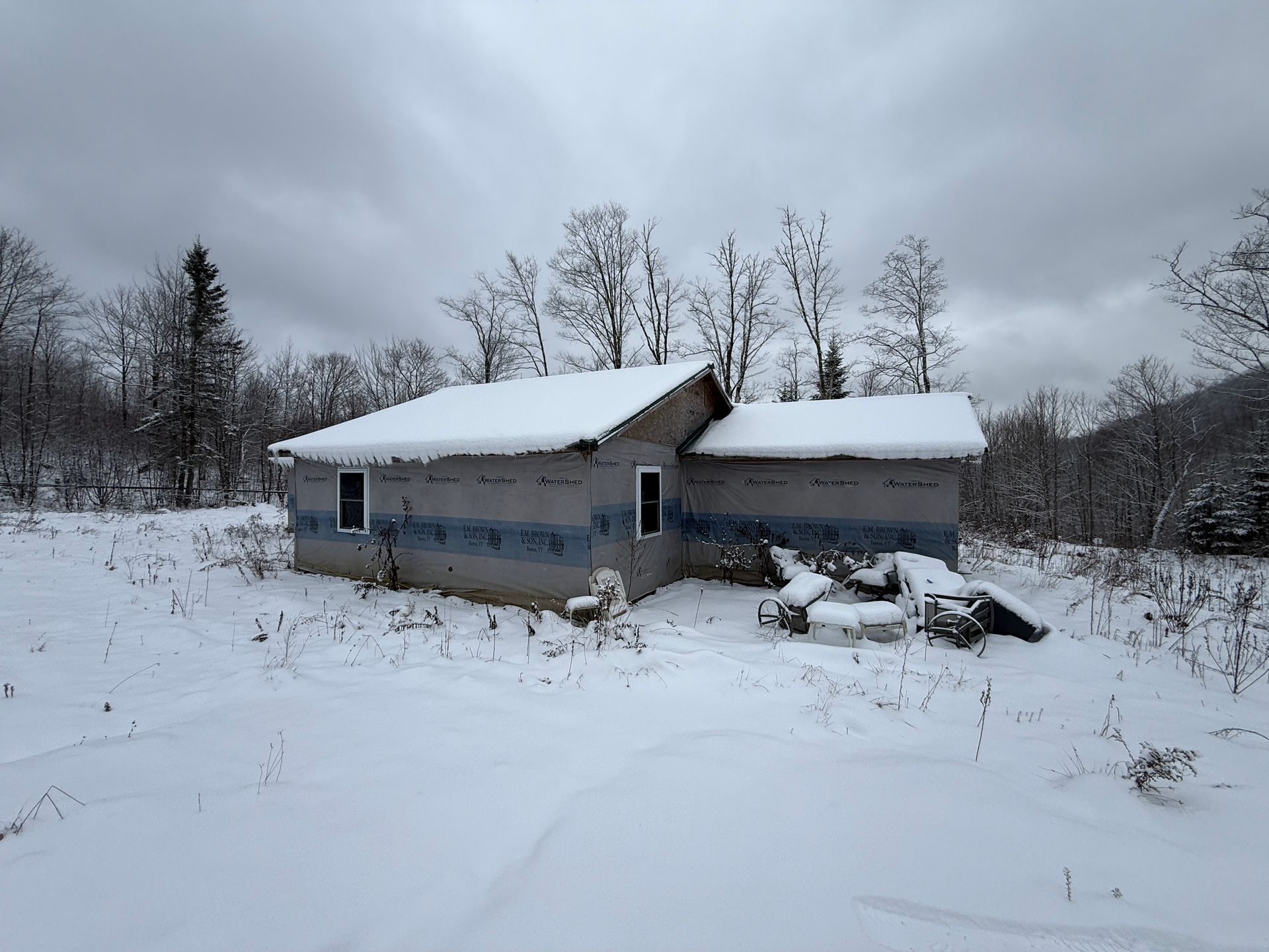Snow-covered small building in a field, bare trees in the background under a cloudy sky.
