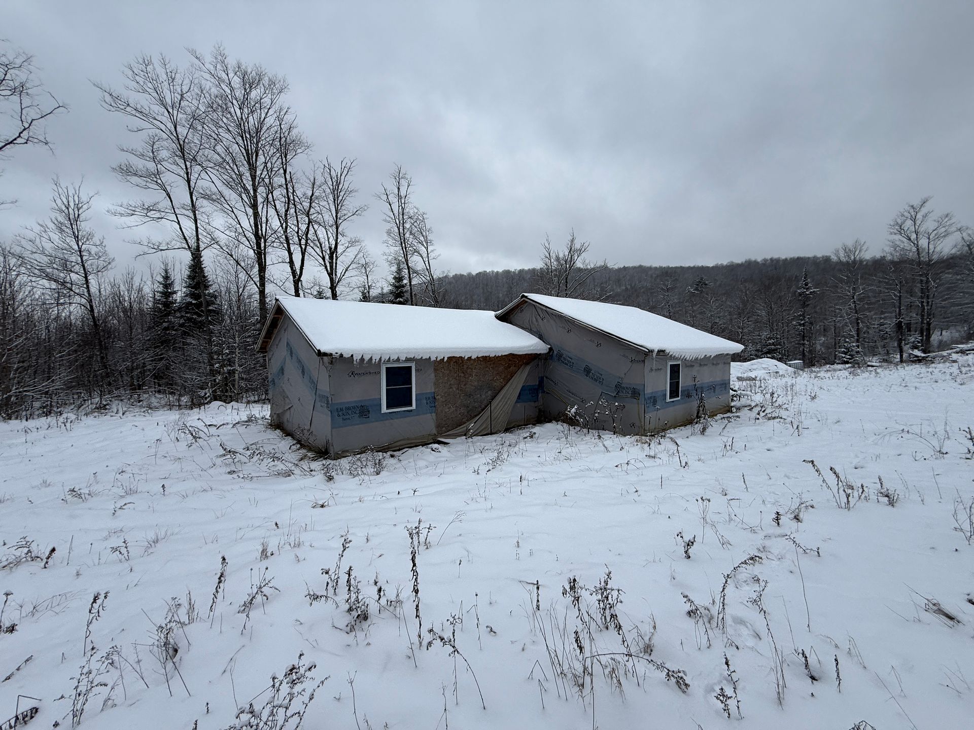 House under construction in a snowy field with bare trees on a cloudy day.