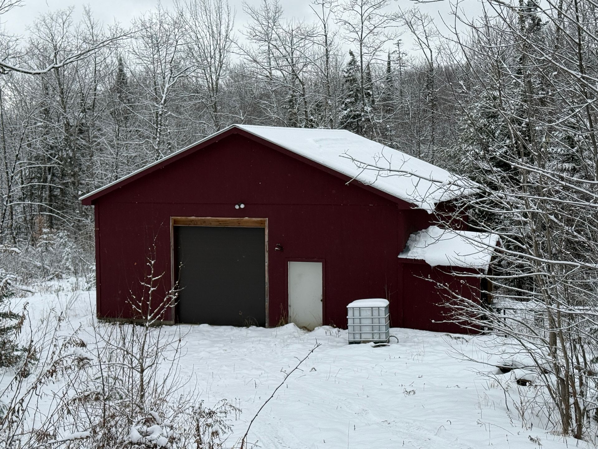 Red building with a garage door and small white door, in a snowy wooded area.