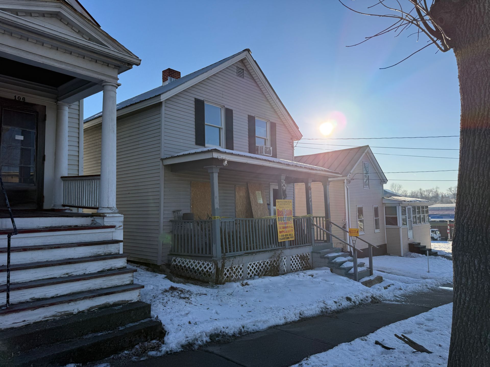 Houses on snowy street with sun shining. The middle house has a porch and boarded-up windows.