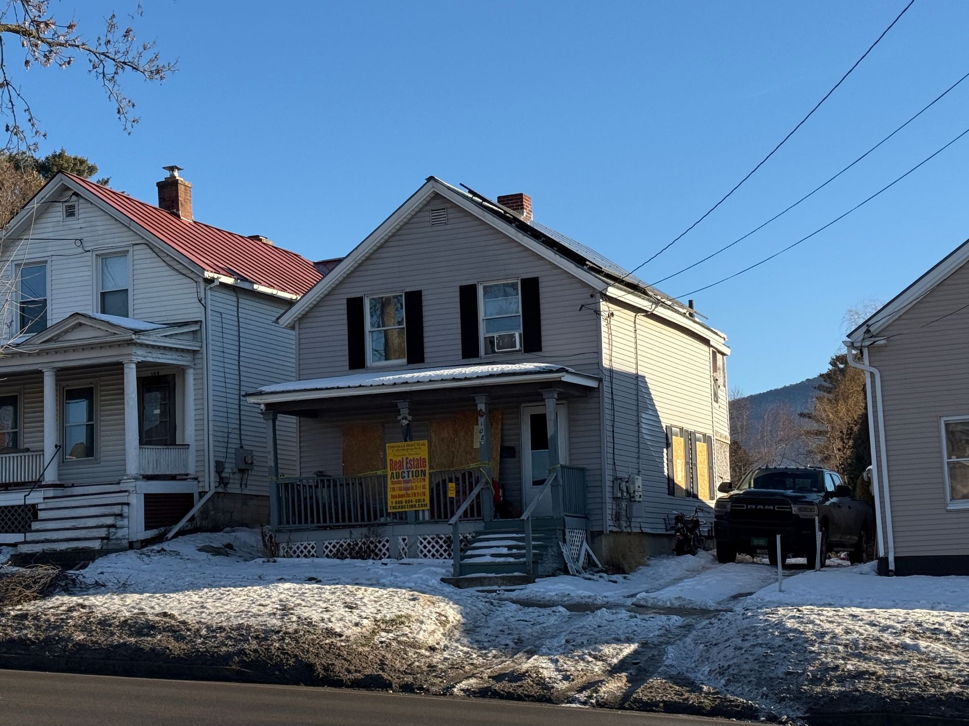 Two-story house with porch, snow-covered ground, parked truck, power lines, and neighboring houses on a sunny day.