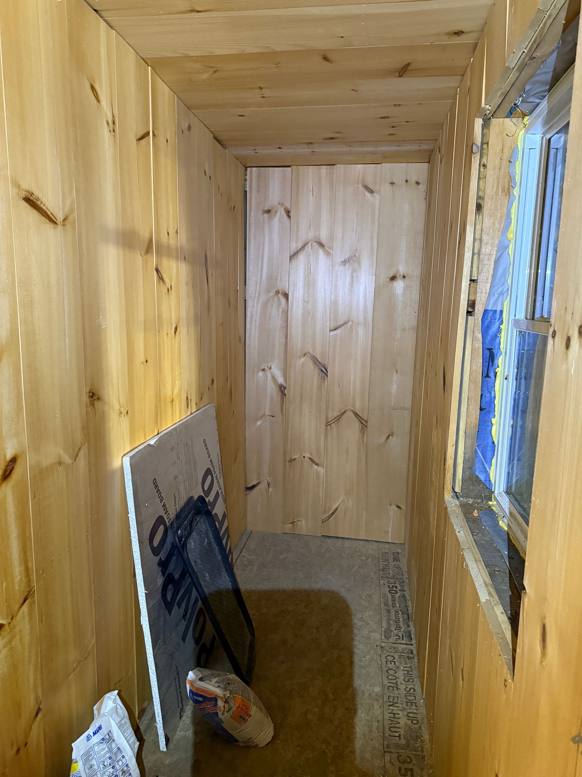Narrow, wood-paneled room with a window on the right. A door is at the end. Materials are on the floor.
