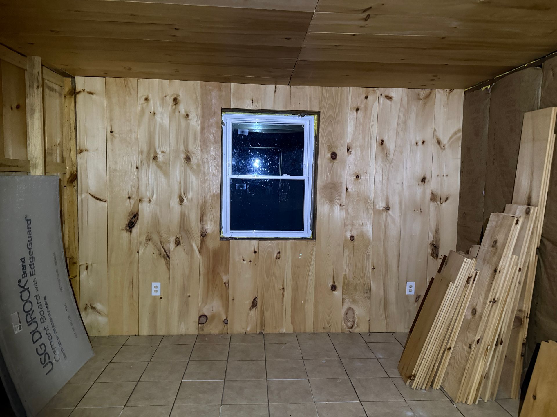 Interior room with wood panel walls, window, and wood planks stacked in the corner.