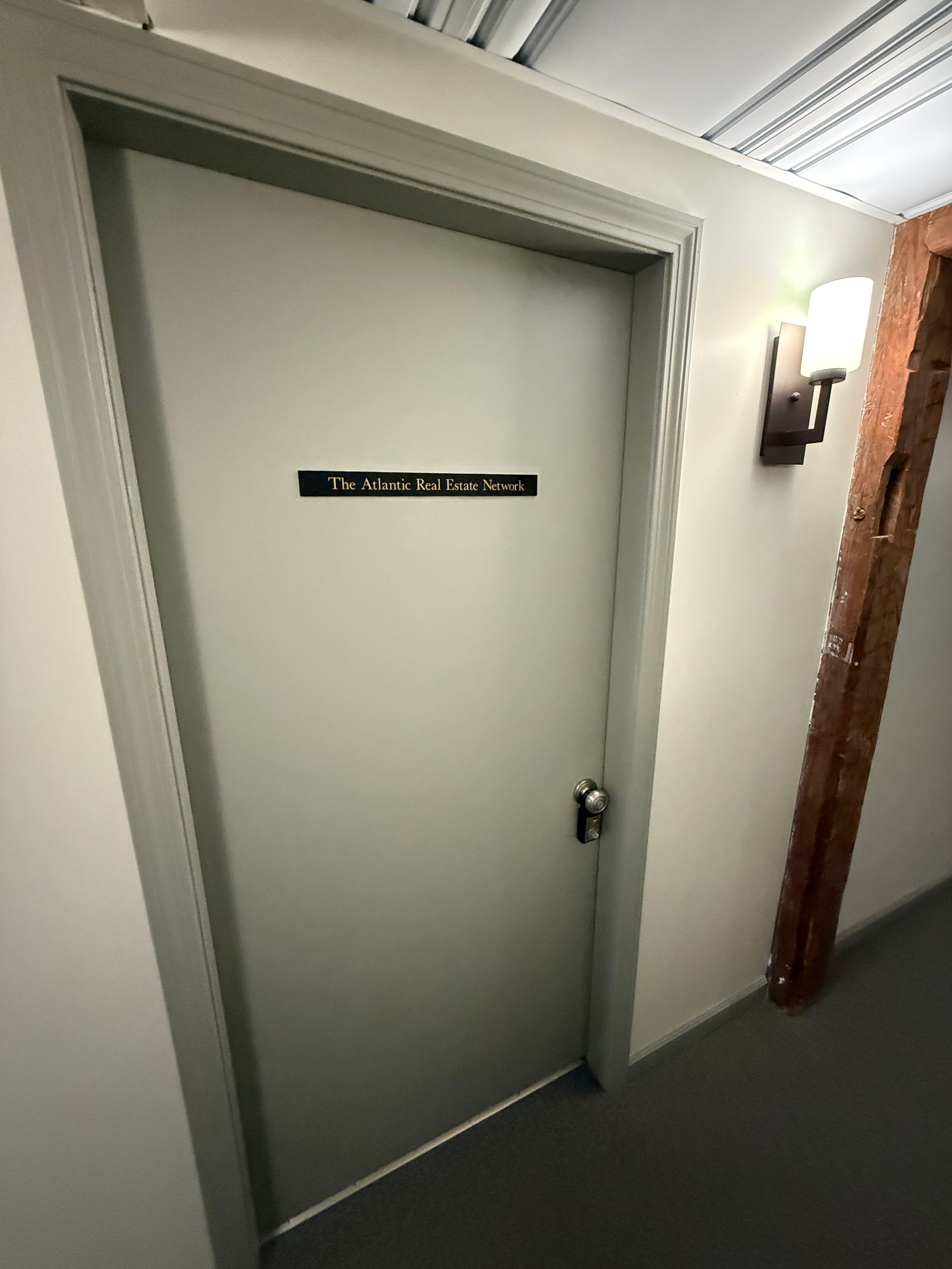 A closed light gray door with a sign, in a hallway. A wall sconce and brown wooden beam are visible.