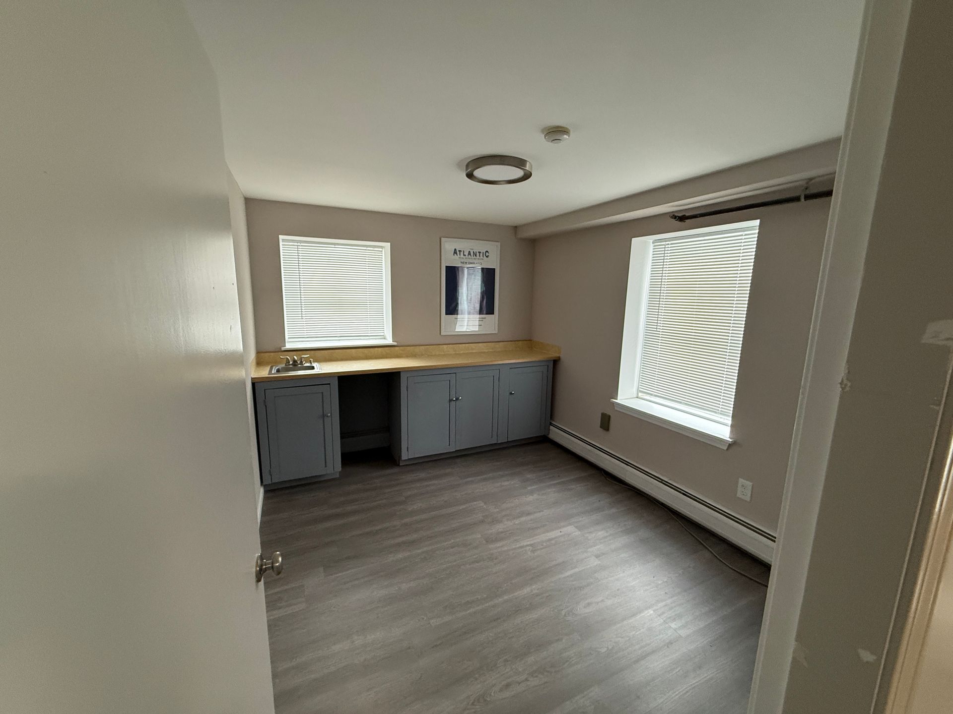 Empty room with gray cabinets, windows with blinds, and wood-look flooring.