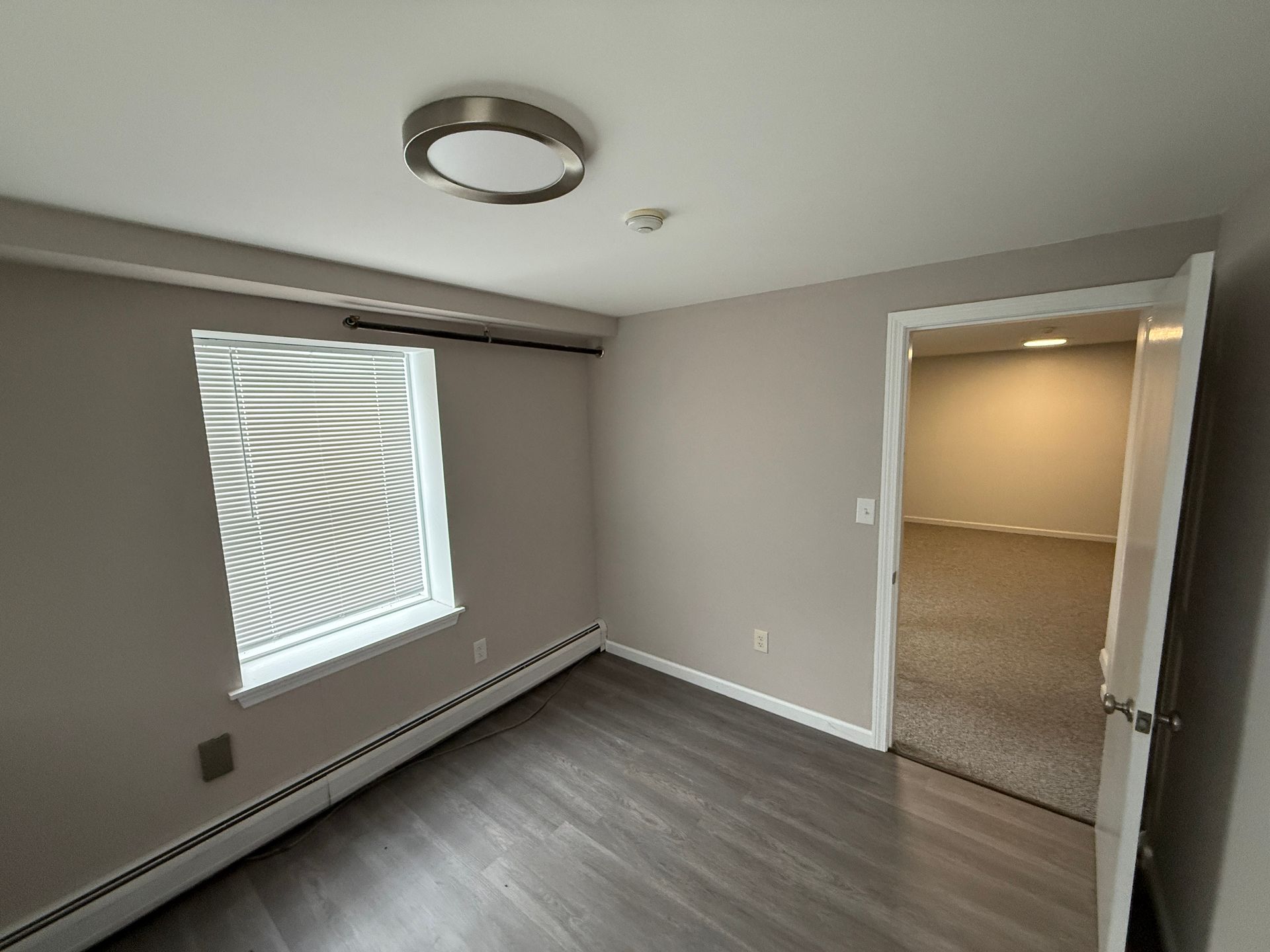 Empty room with wood-look flooring, a window with blinds, and an open doorway leading to another room.