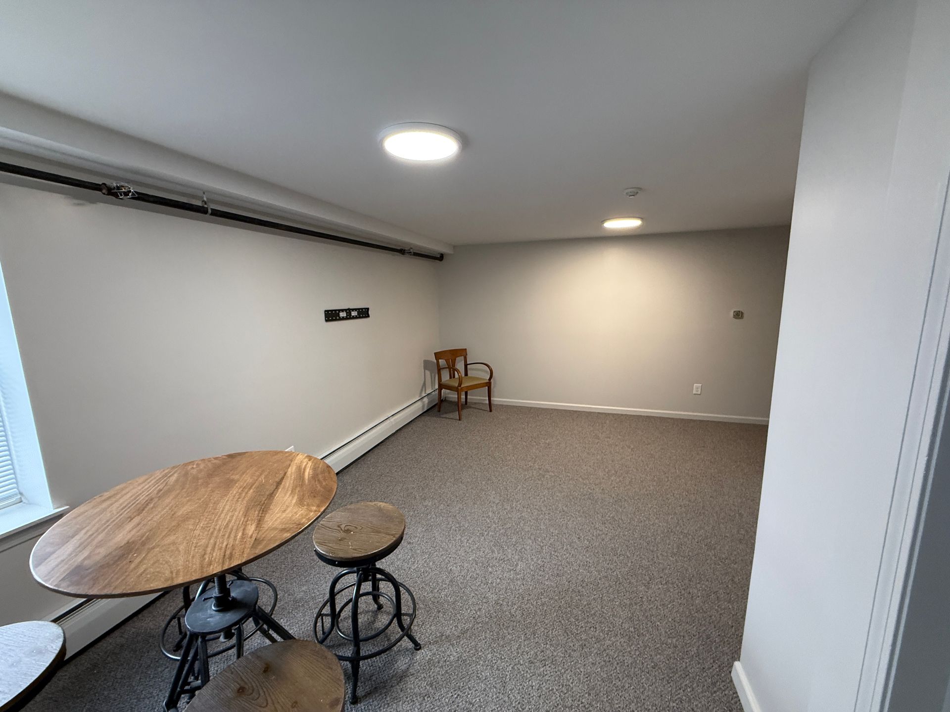 Empty room with a round wooden table, two stools, and a chair. Gray carpet, white walls.