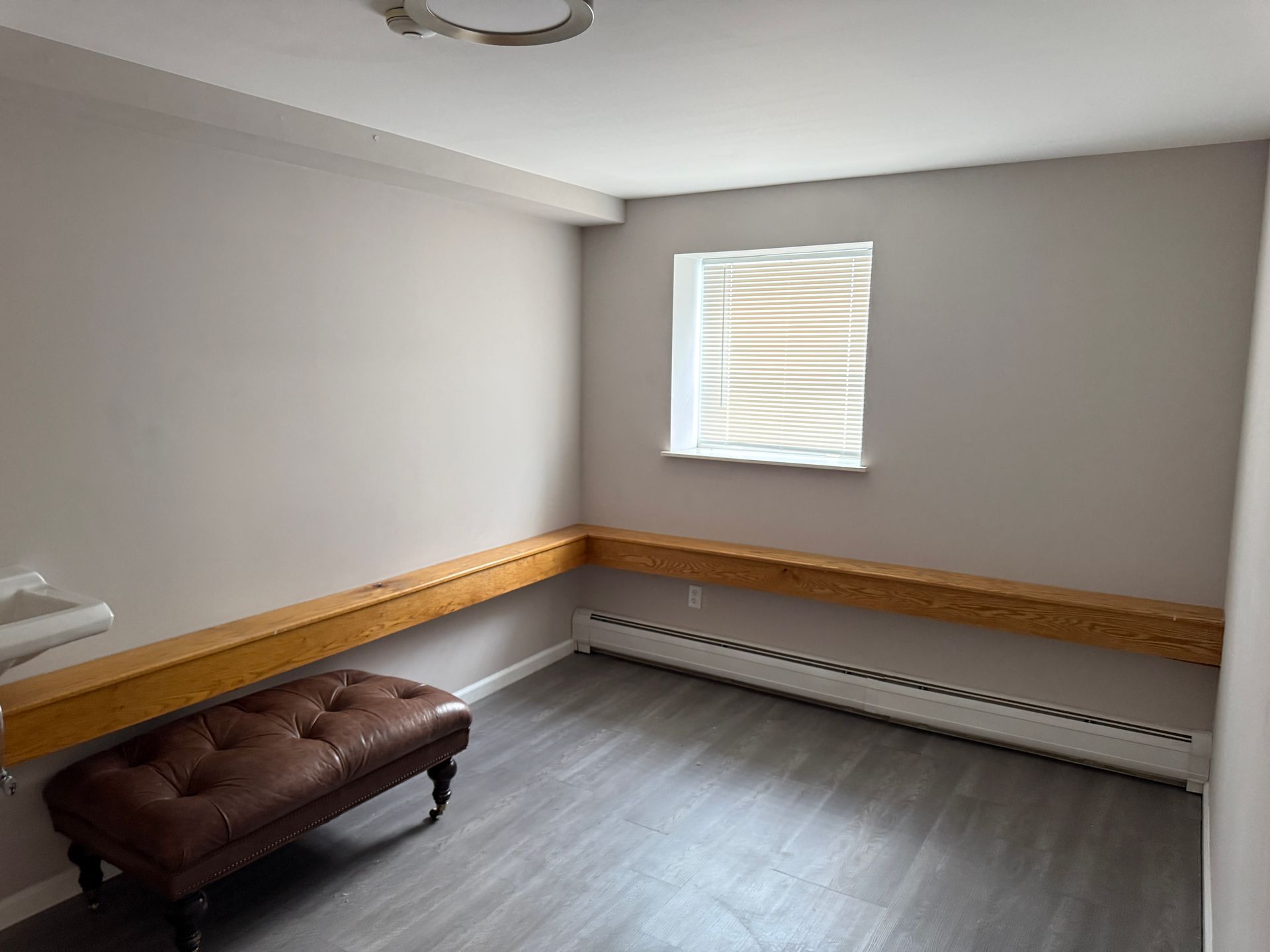 Empty room with gray walls, wood shelf, brown ottoman, window with blinds.