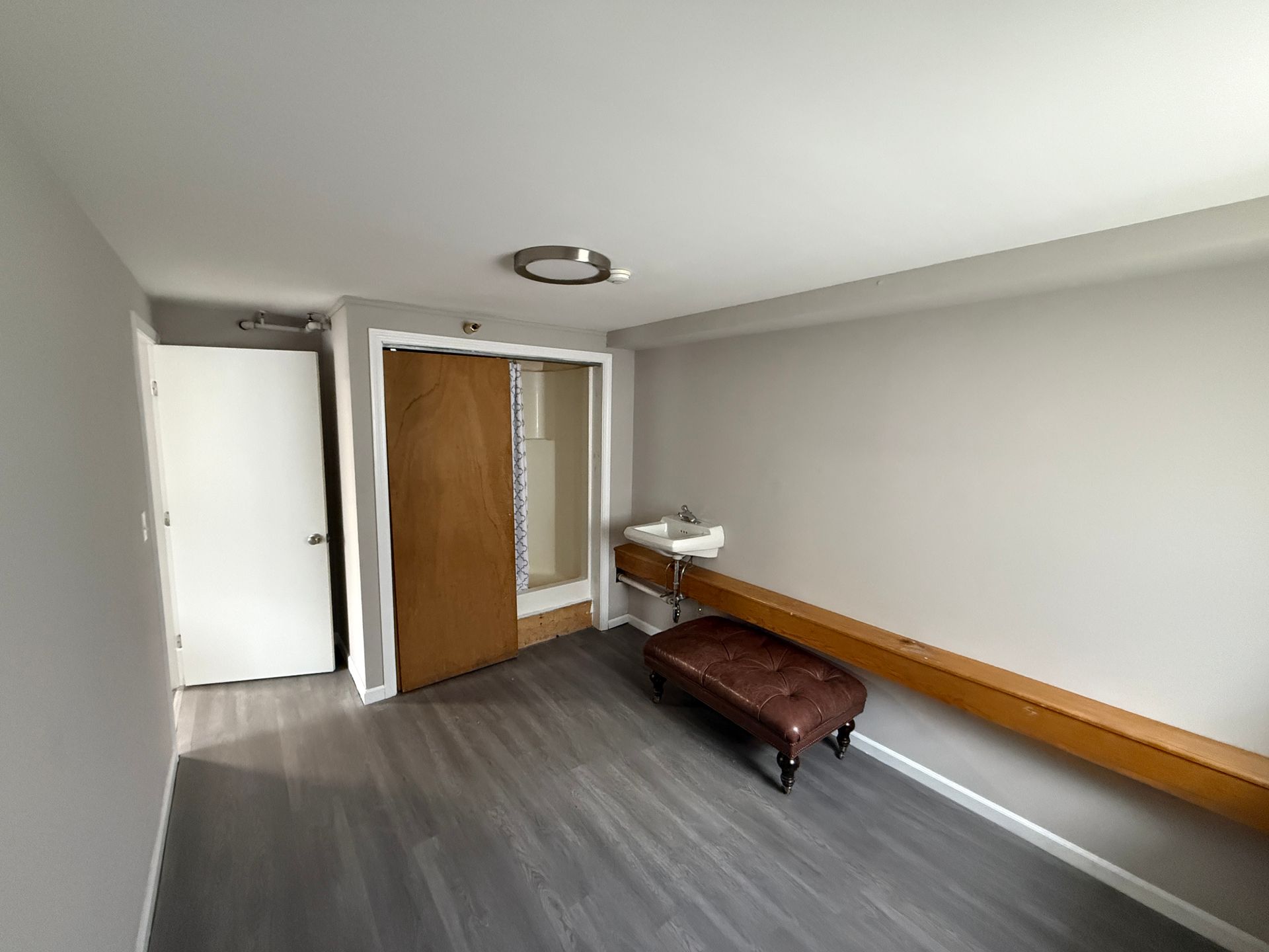 Empty room with gray walls, wood floor, small sink, door, and bench.