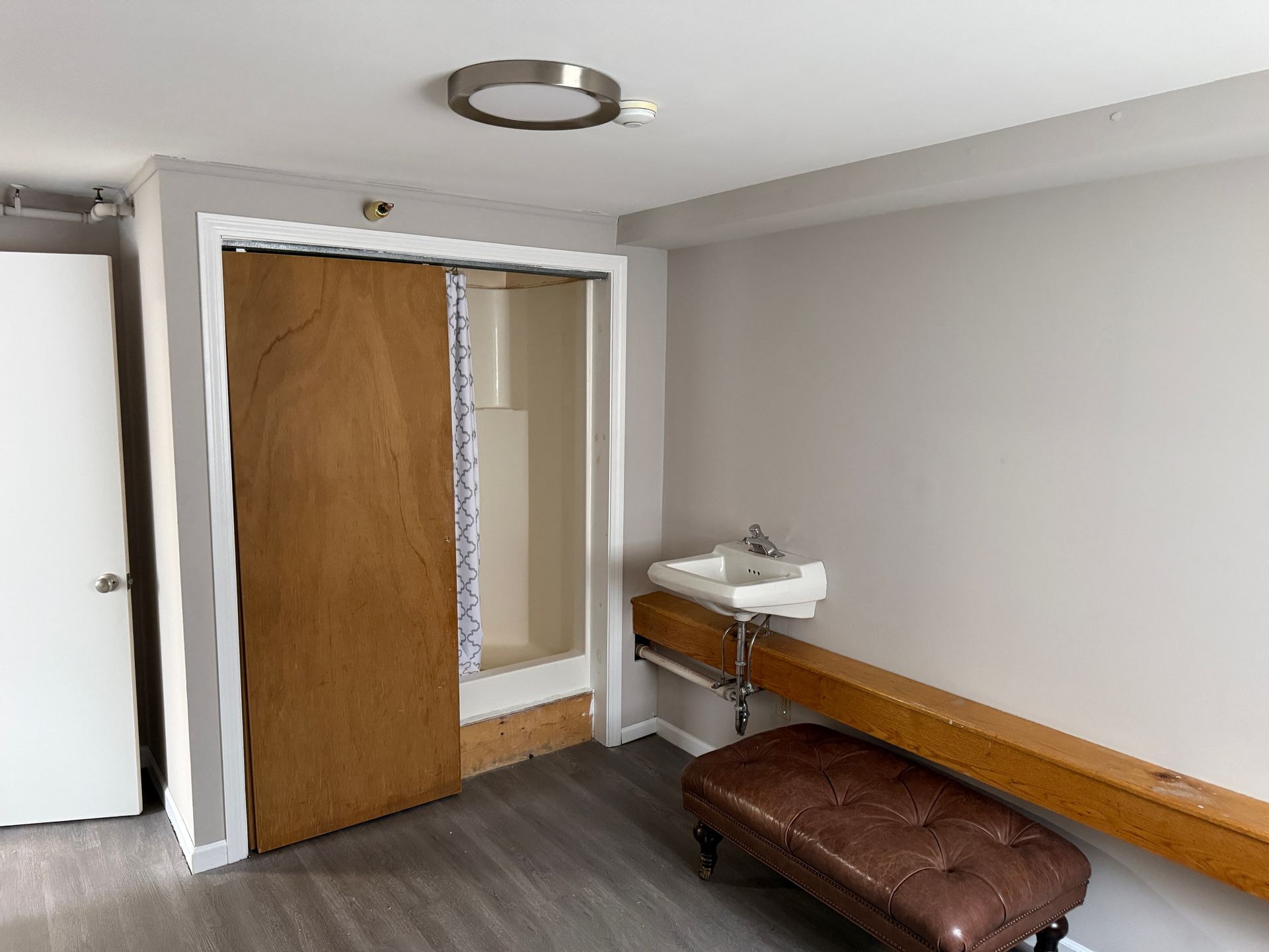 A small room with a sink, wooden cabinet, and stool; gray walls, wood-look flooring, and a round ceiling light.