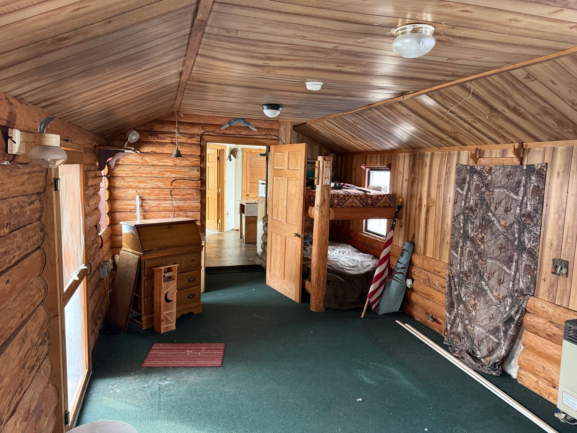 Rustic interior of a cabin with log walls, bunk beds, and a doorway leading to another room. Green carpet.