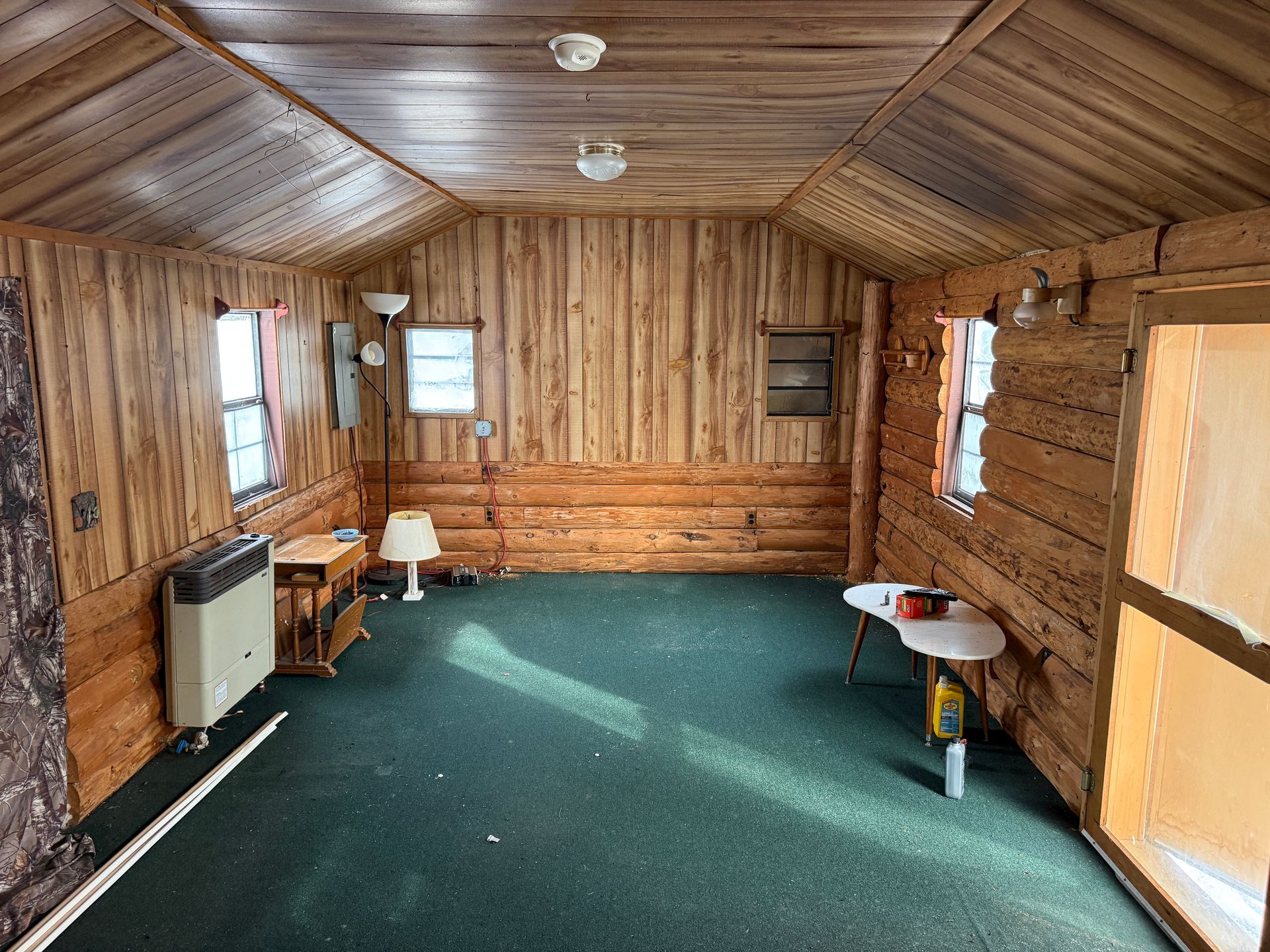 Interior of a cabin with wood-paneled walls and ceiling. Green carpet, small windows, and simple furnishings.