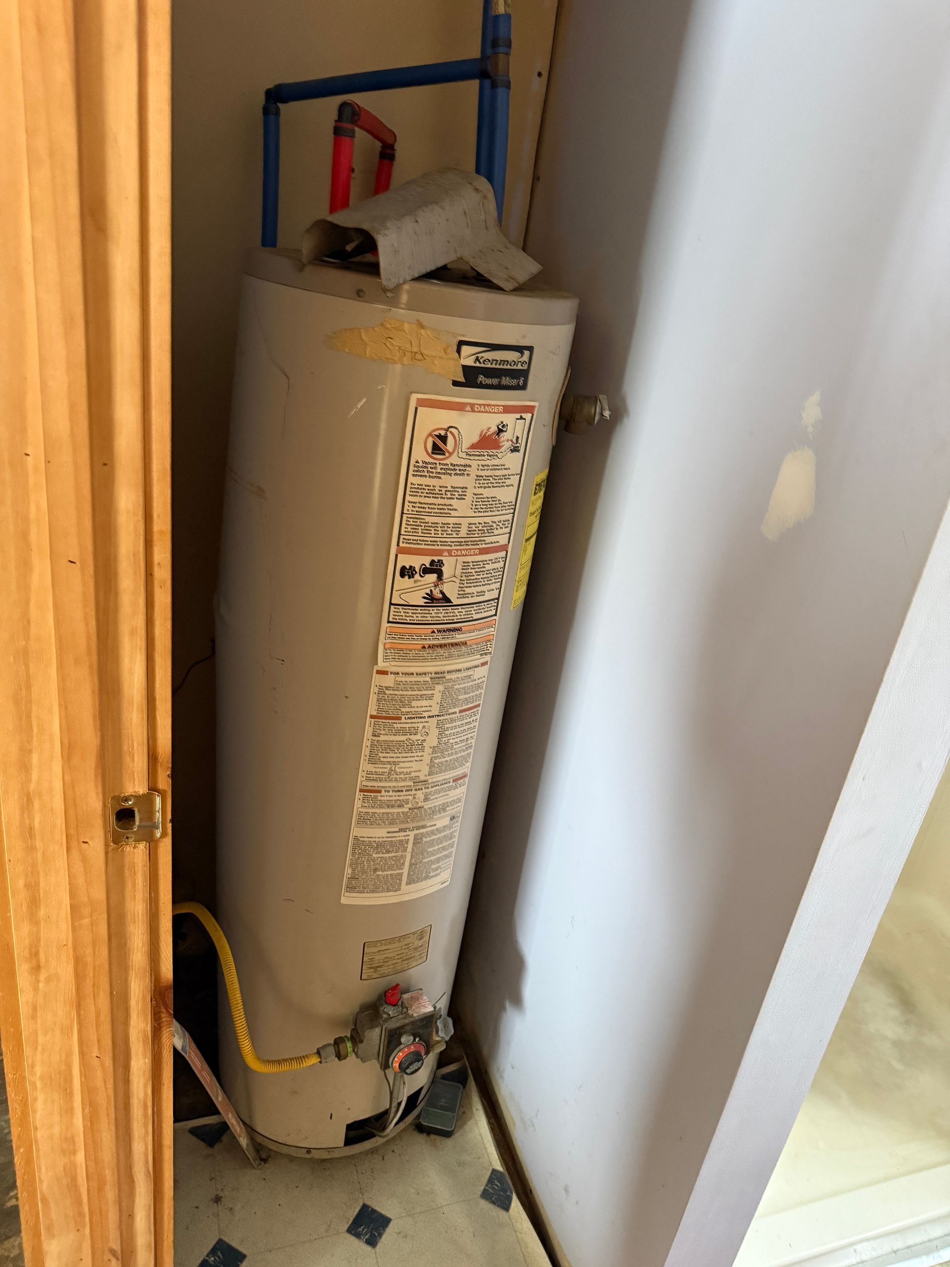 A water heater in a narrow closet, tilting slightly. Gray tank with red and yellow pipes.