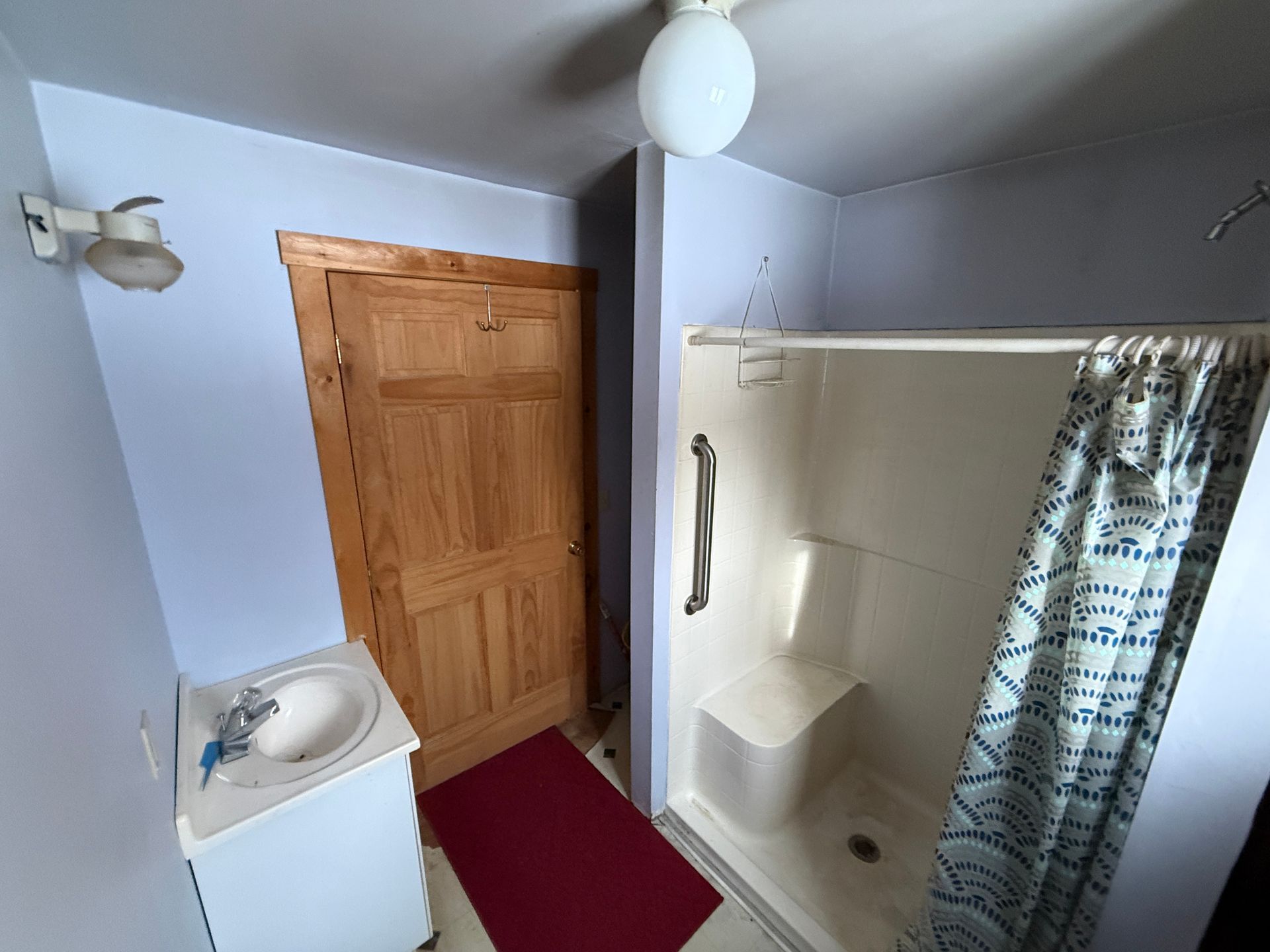 Bathroom with a white sink, shower, and light blue walls. Red rug in front of wooden door.