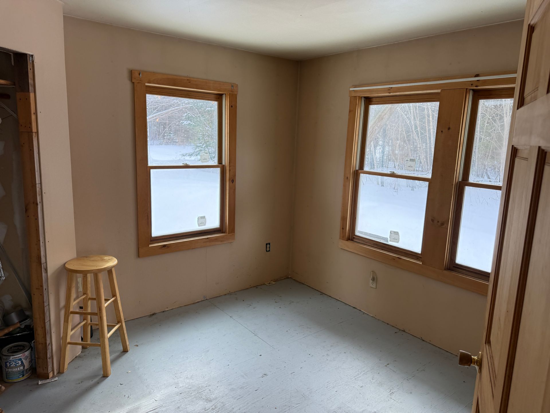 Empty room with two windows, stool, and door. Walls are beige, windows have brown frames.