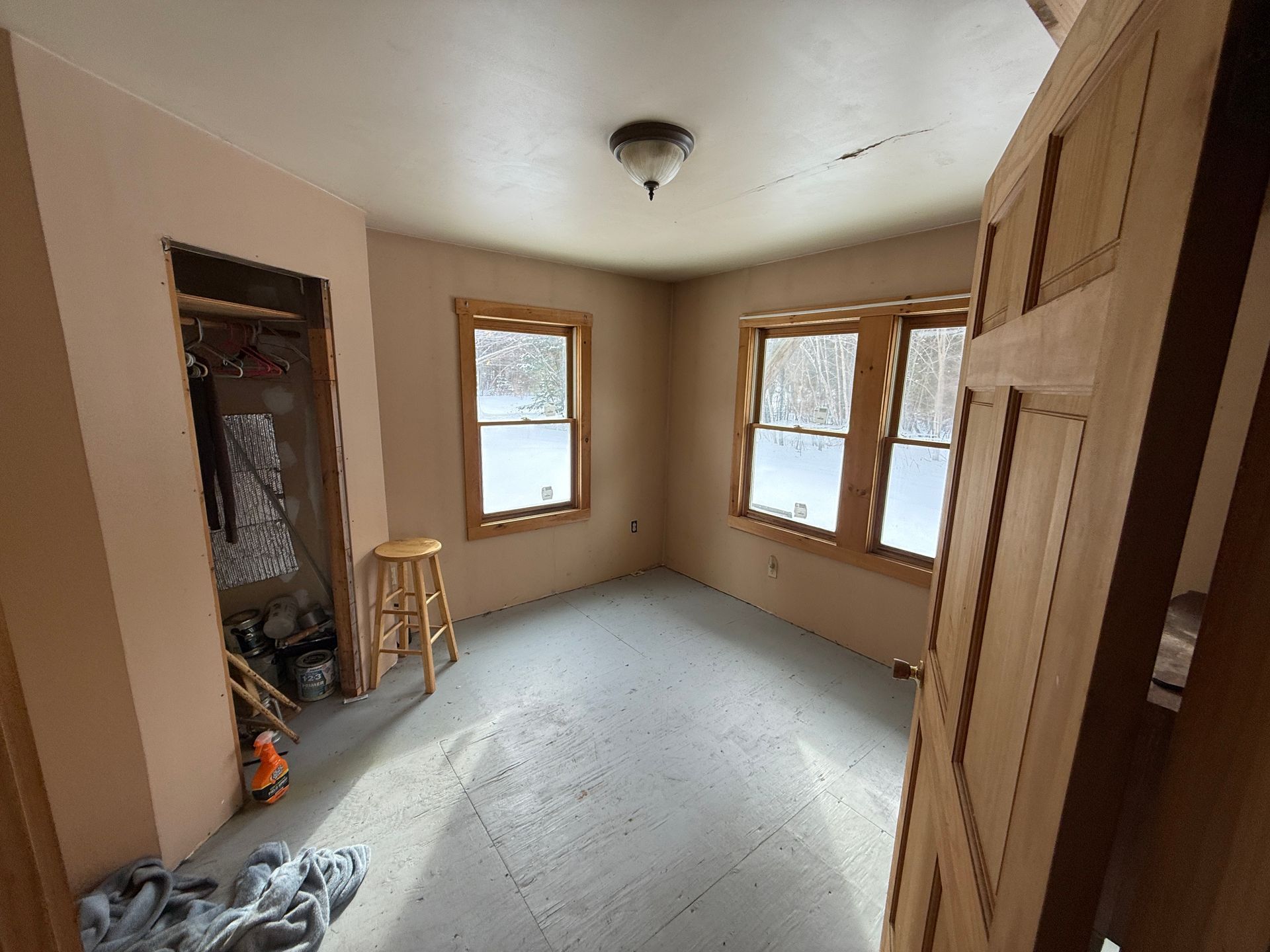 Empty room with two windows, closet, and a partially open door; beige walls and floor, natural wood window frames.