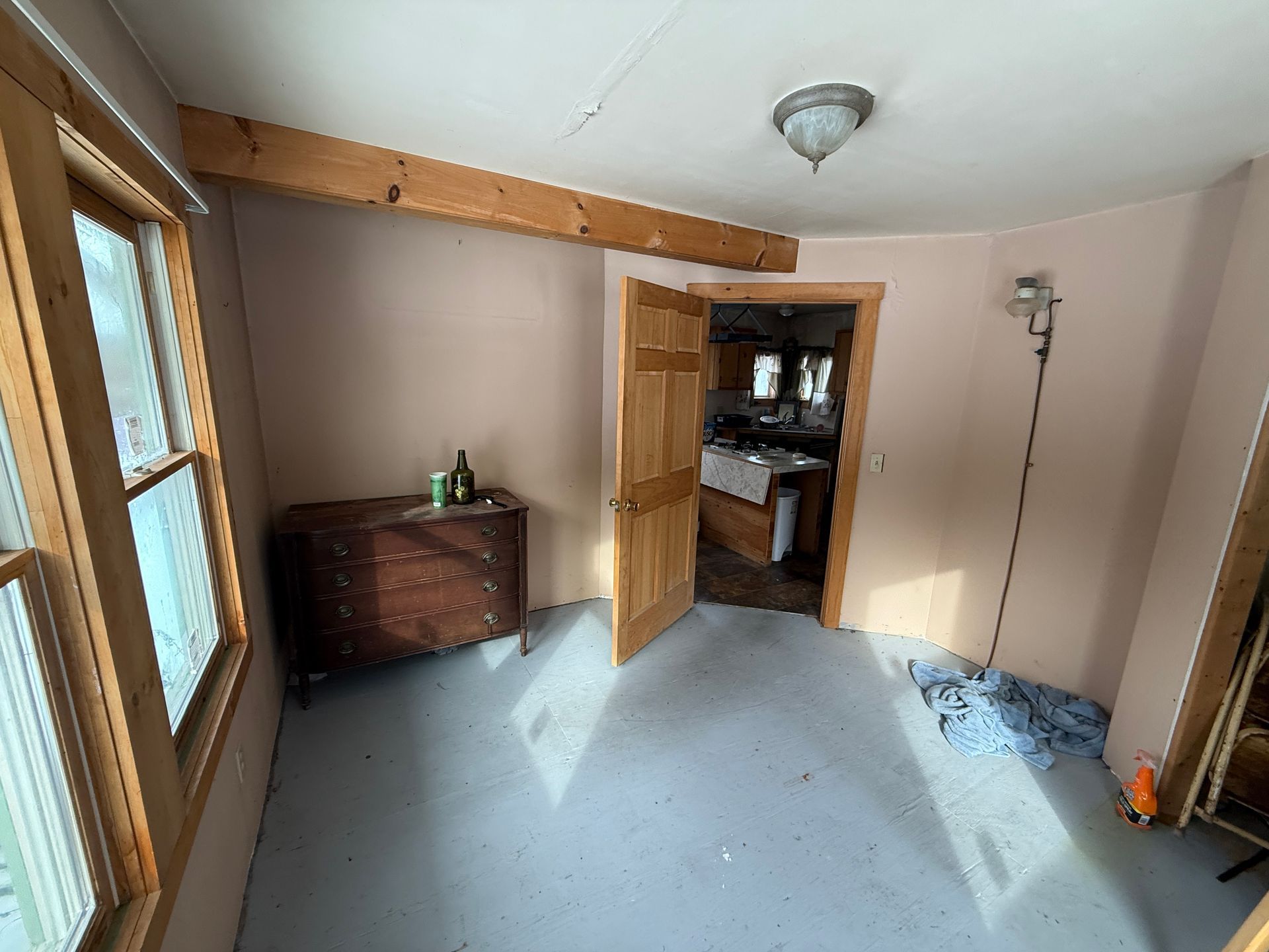Small, empty room with open door to a kitchen, window on the left, and a dresser.