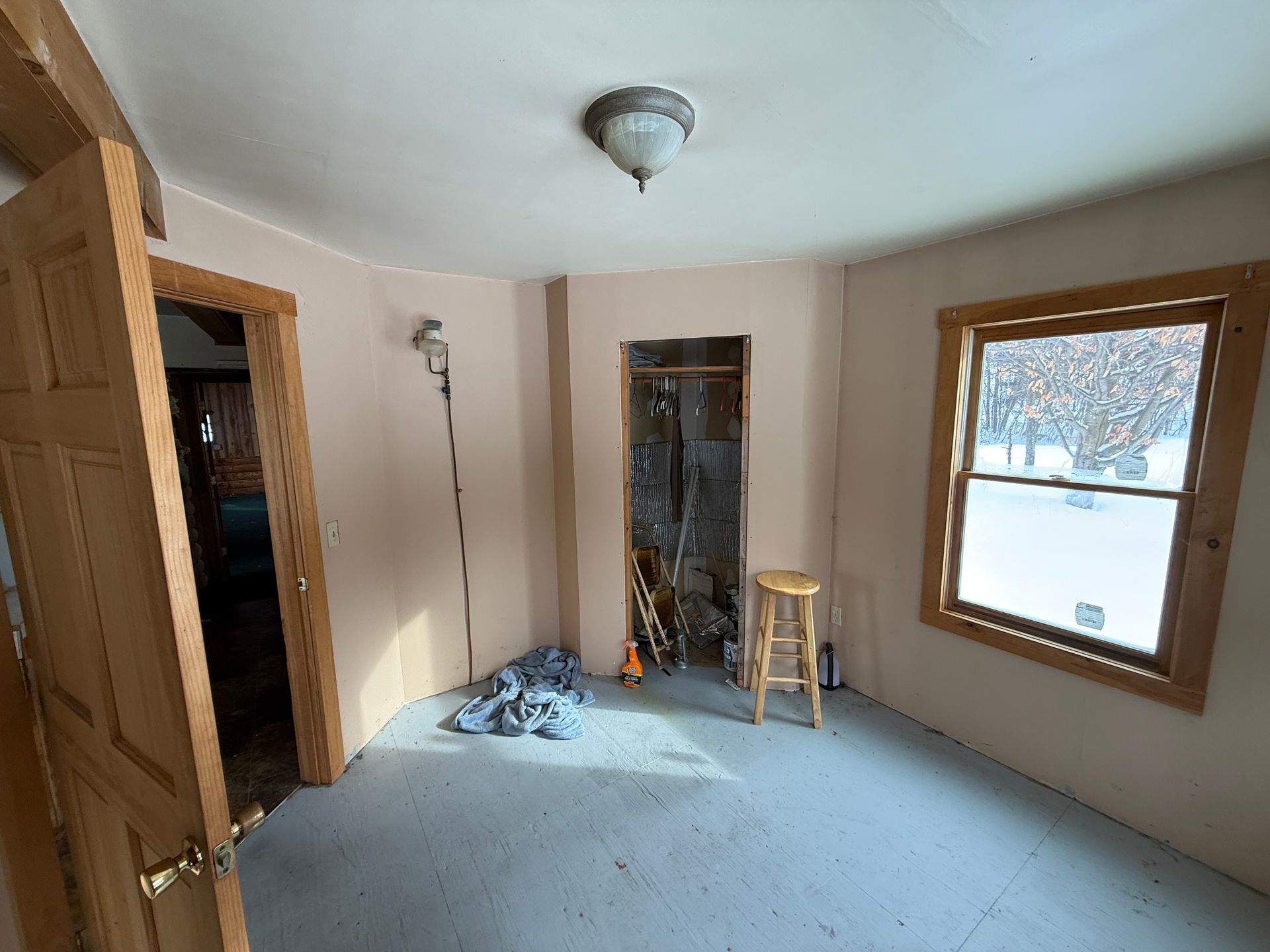 Empty room with open door, closet space, and window. Pink walls, gray floor, and a wooden stool.