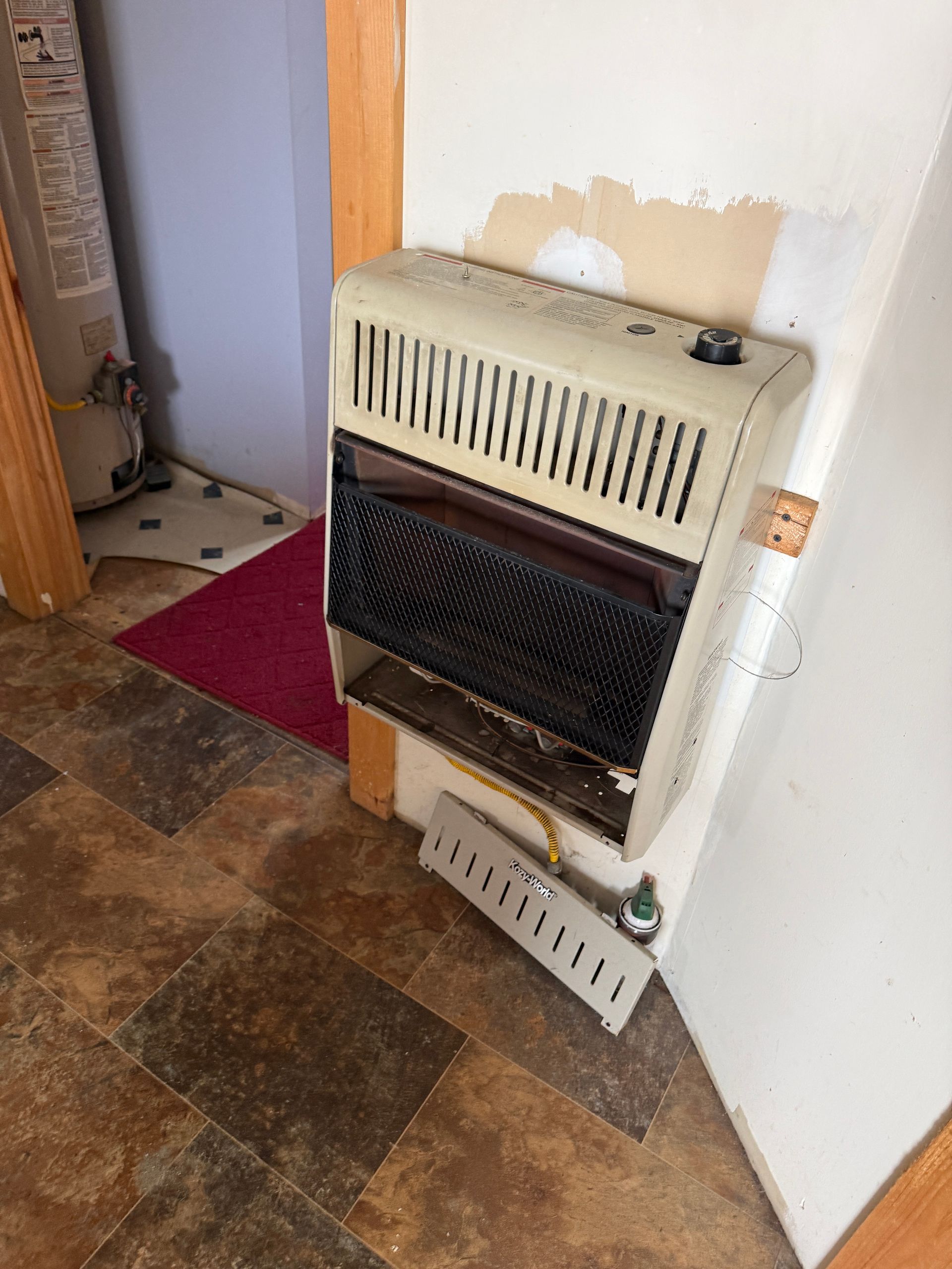 A wall-mounted gas heater in a corner, next to a door and brown flooring.