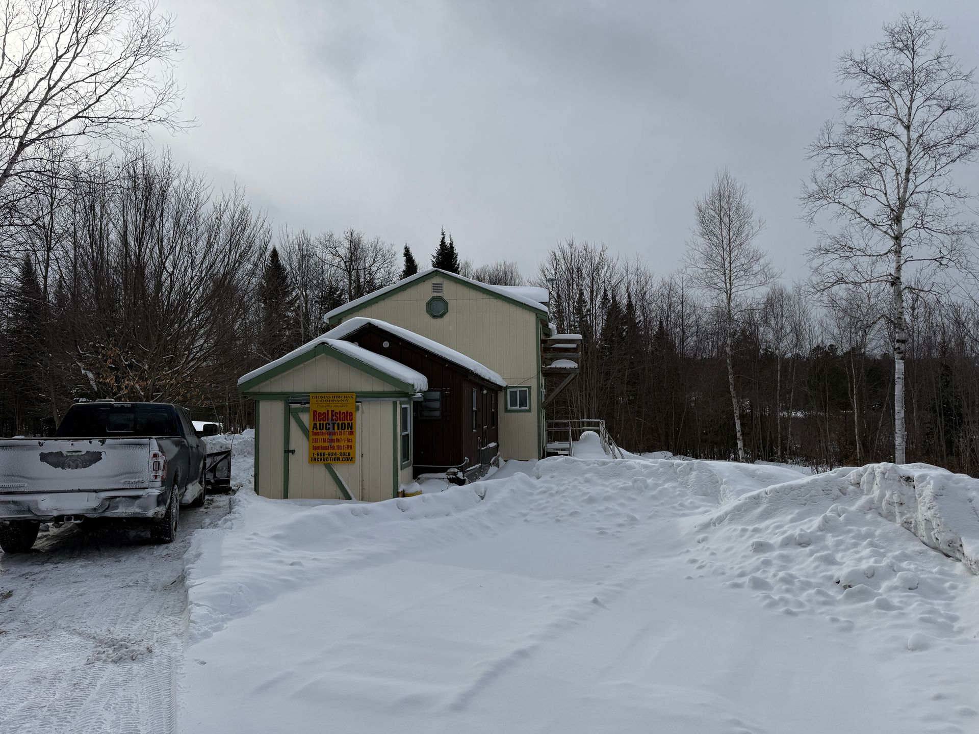 Snow-covered lot with a two-story building and truck. Trees in background, overcast sky.