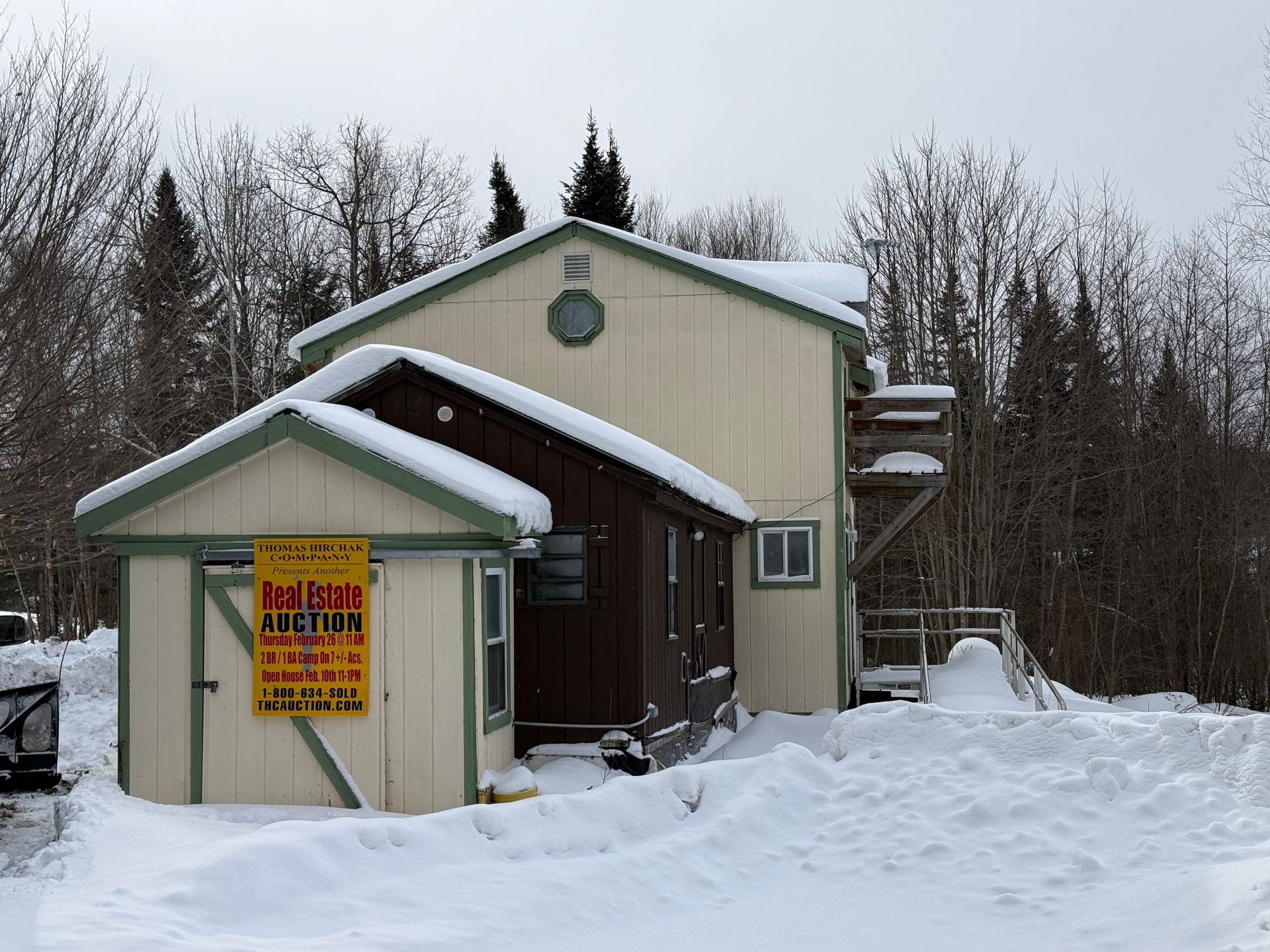 A snow-covered building with a real estate auction sign. A wooden shed is attached.