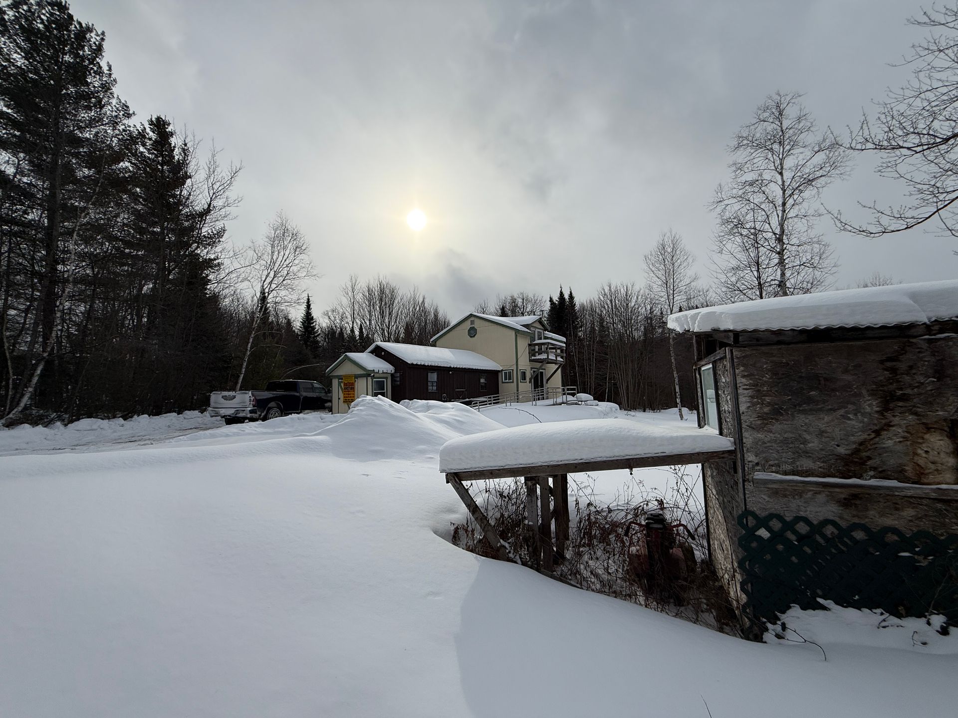 Snow-covered landscape with a house in the background and a partially covered structure in the foreground under a cloudy sky.