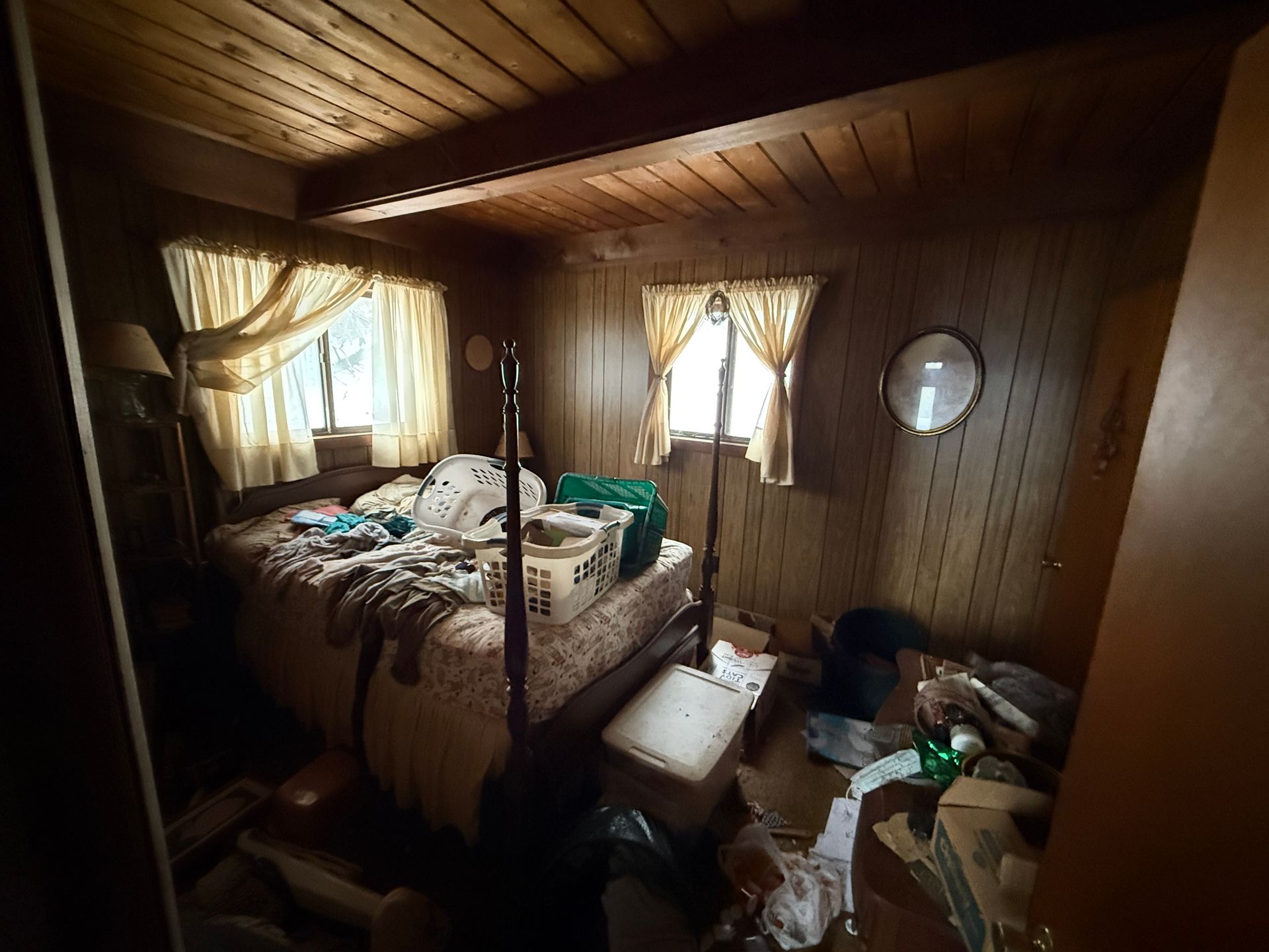 Bedroom: bed with bedding, two windows with curtains, wooden paneling.