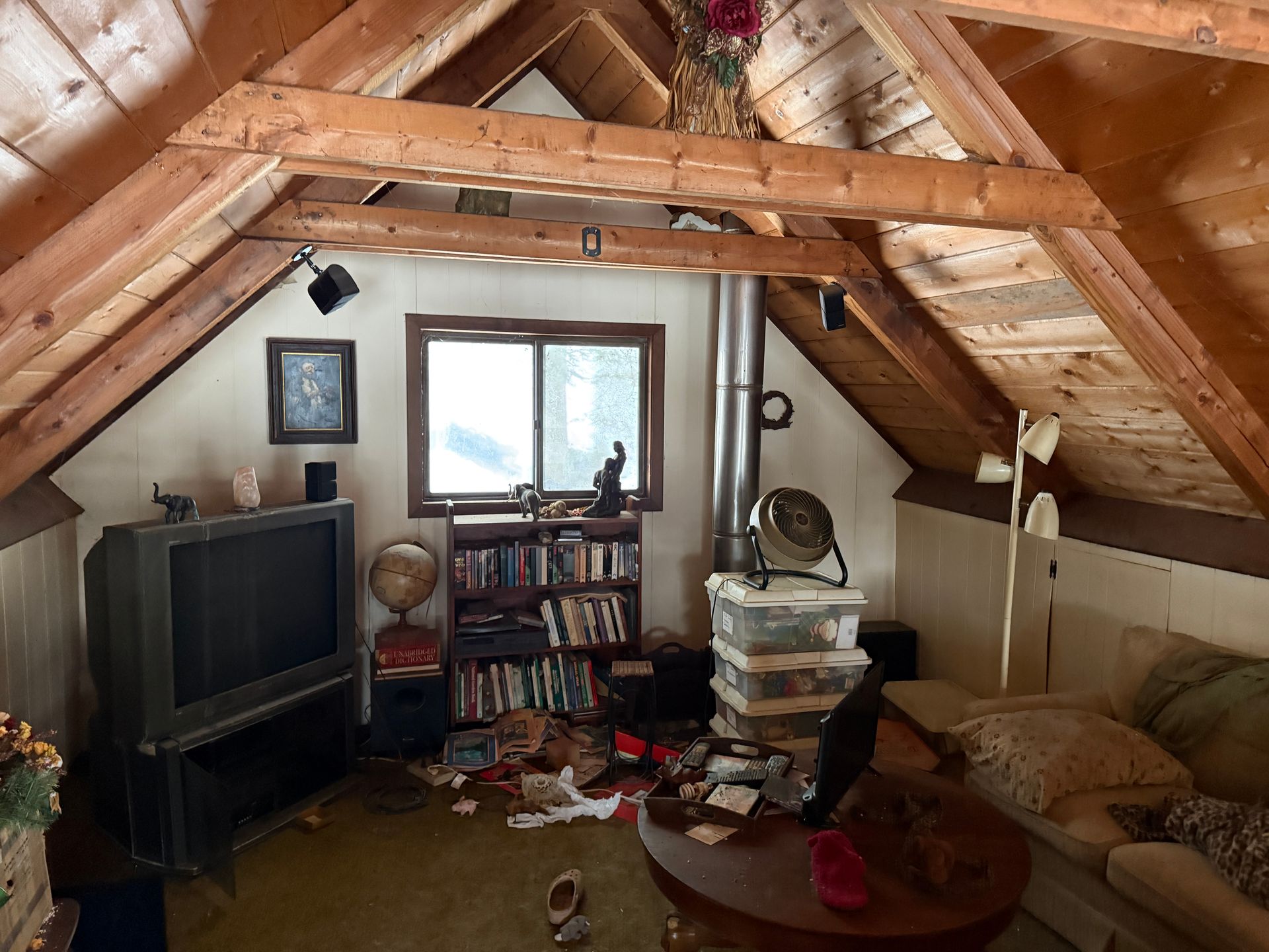 Attic interior with sloped ceiling, wood beams, window, bookshelf, couch, TV.