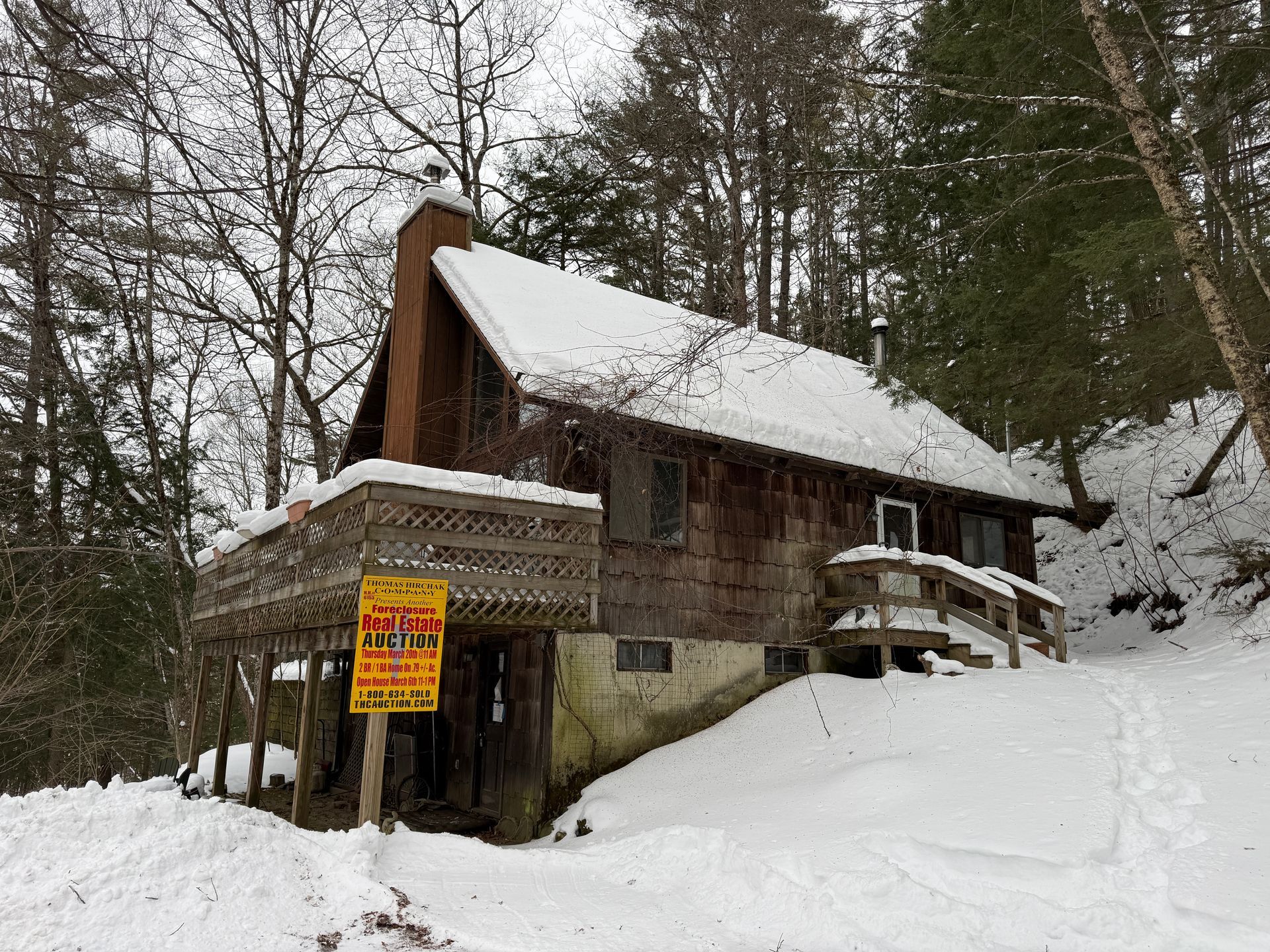 Snow-covered cabin in a wooded area with a deck and a for-sale sign.