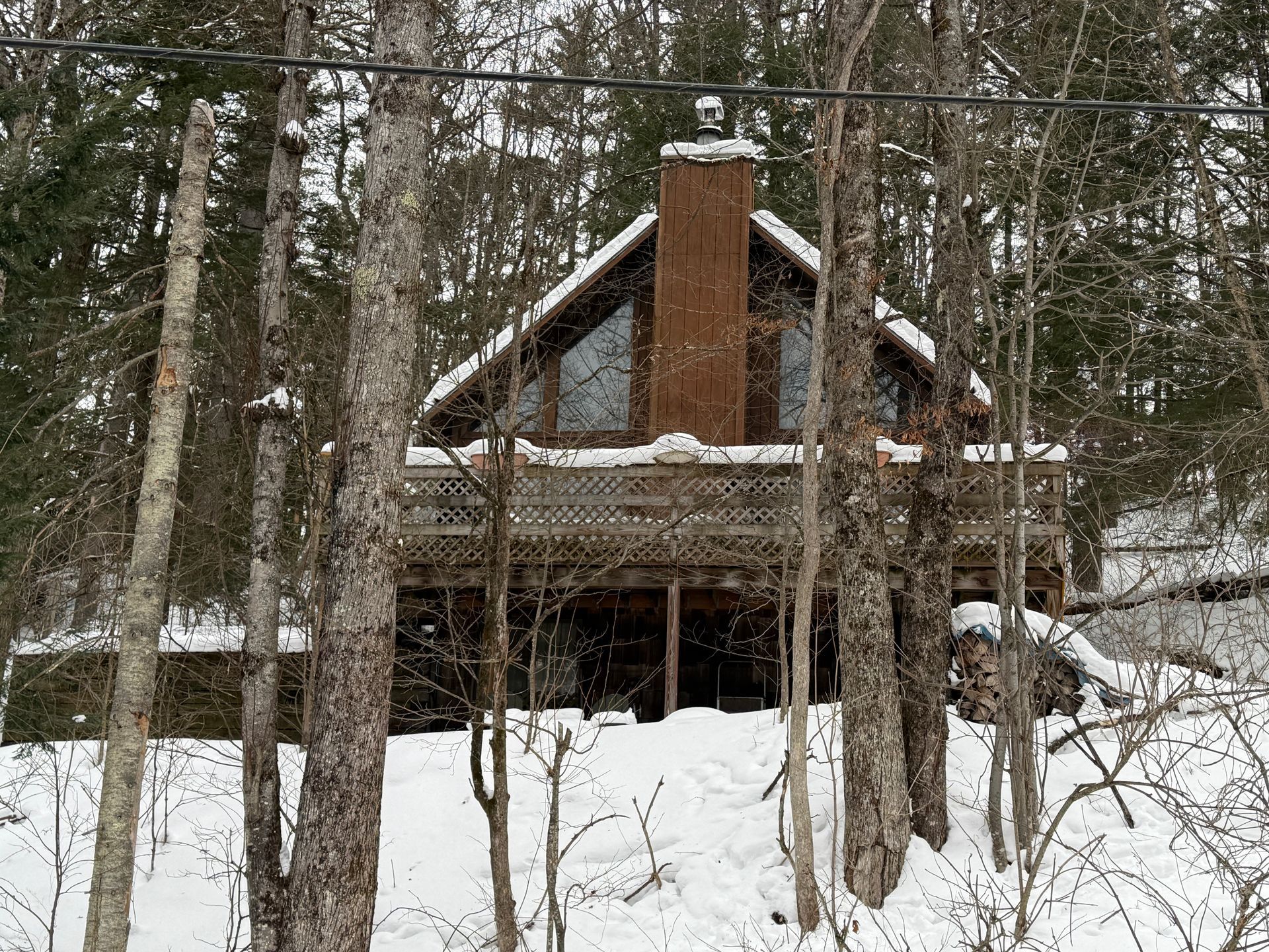 Snowy cabin in the woods, with a deck and chimney.