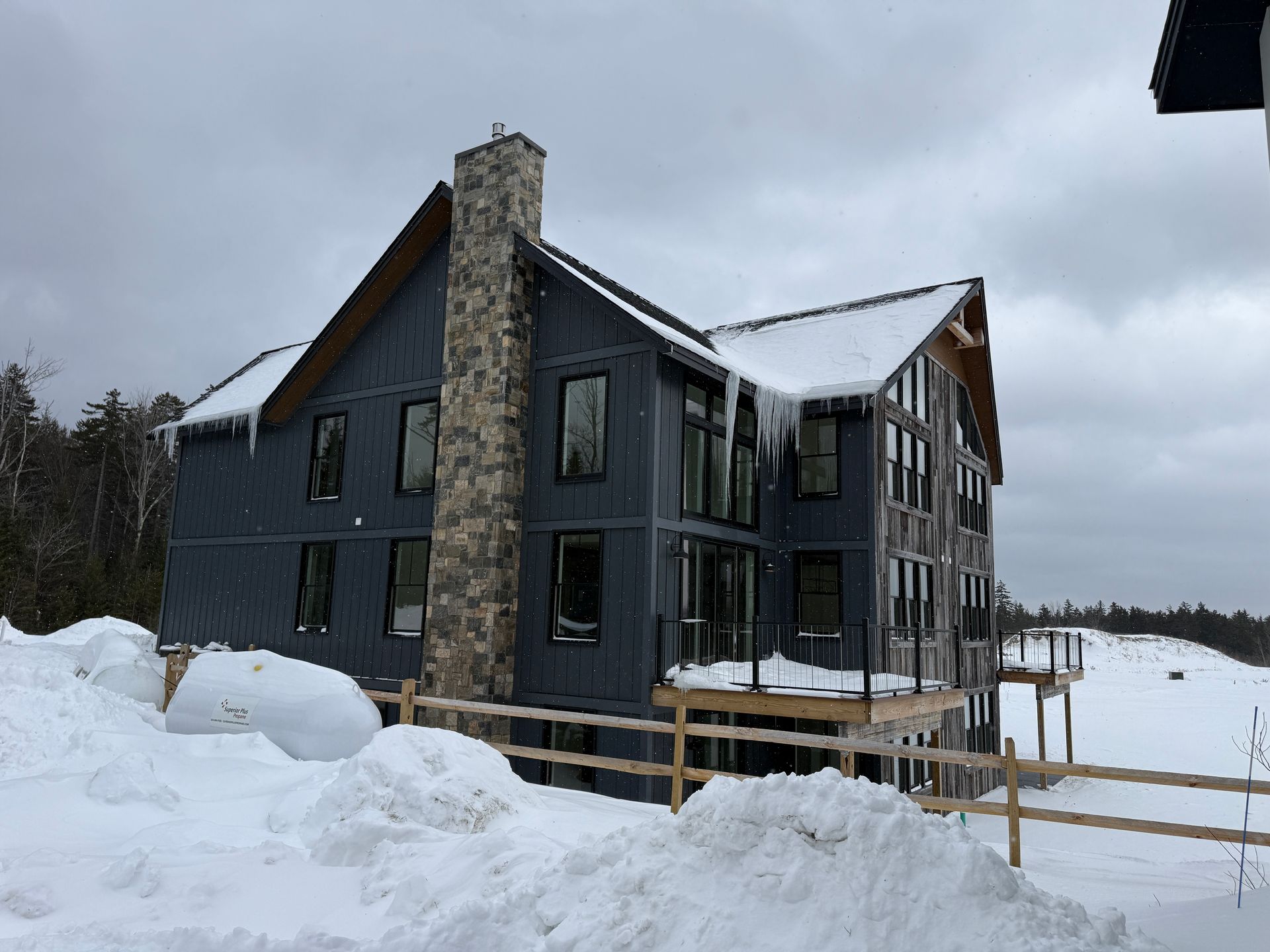 Dark blue cabin covered in snow with icicles, tall stone chimney, wooden deck. Boulder Ridge Foreclosure Property.