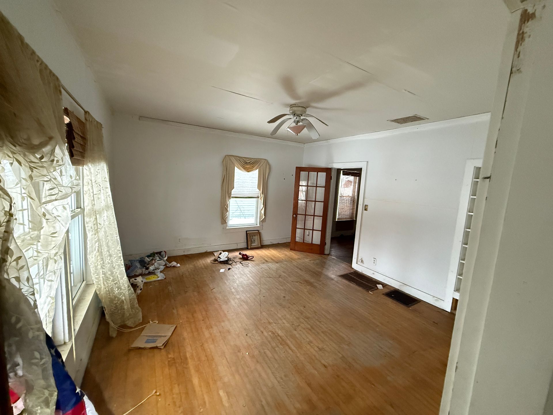 A room with hardwood floors, white walls, a ceiling fan, a window with curtains, and a glass-paneled wooden door.