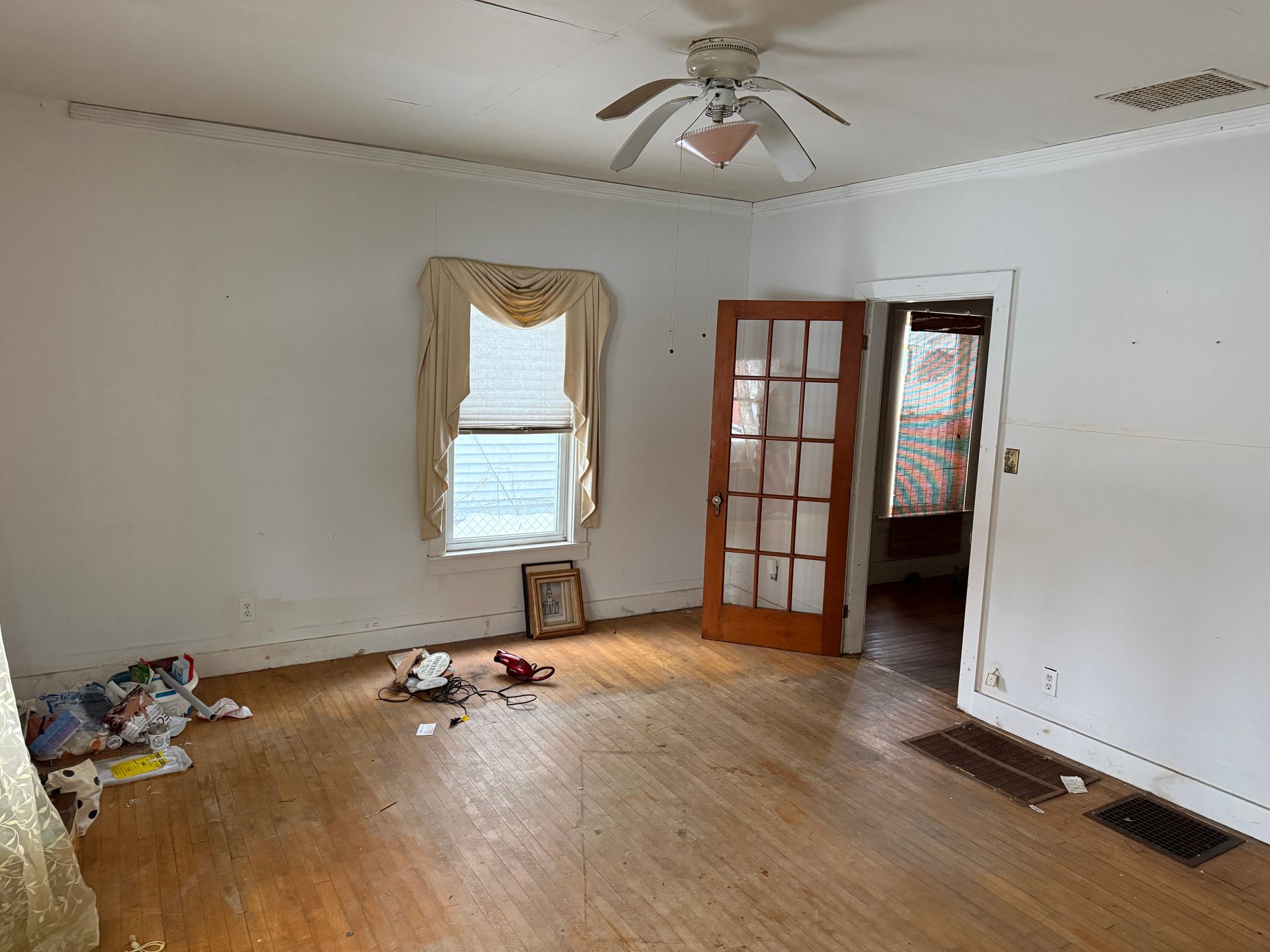 Empty room with hardwood floors, a white ceiling fan, a window with beige curtains, and a wood-framed door.