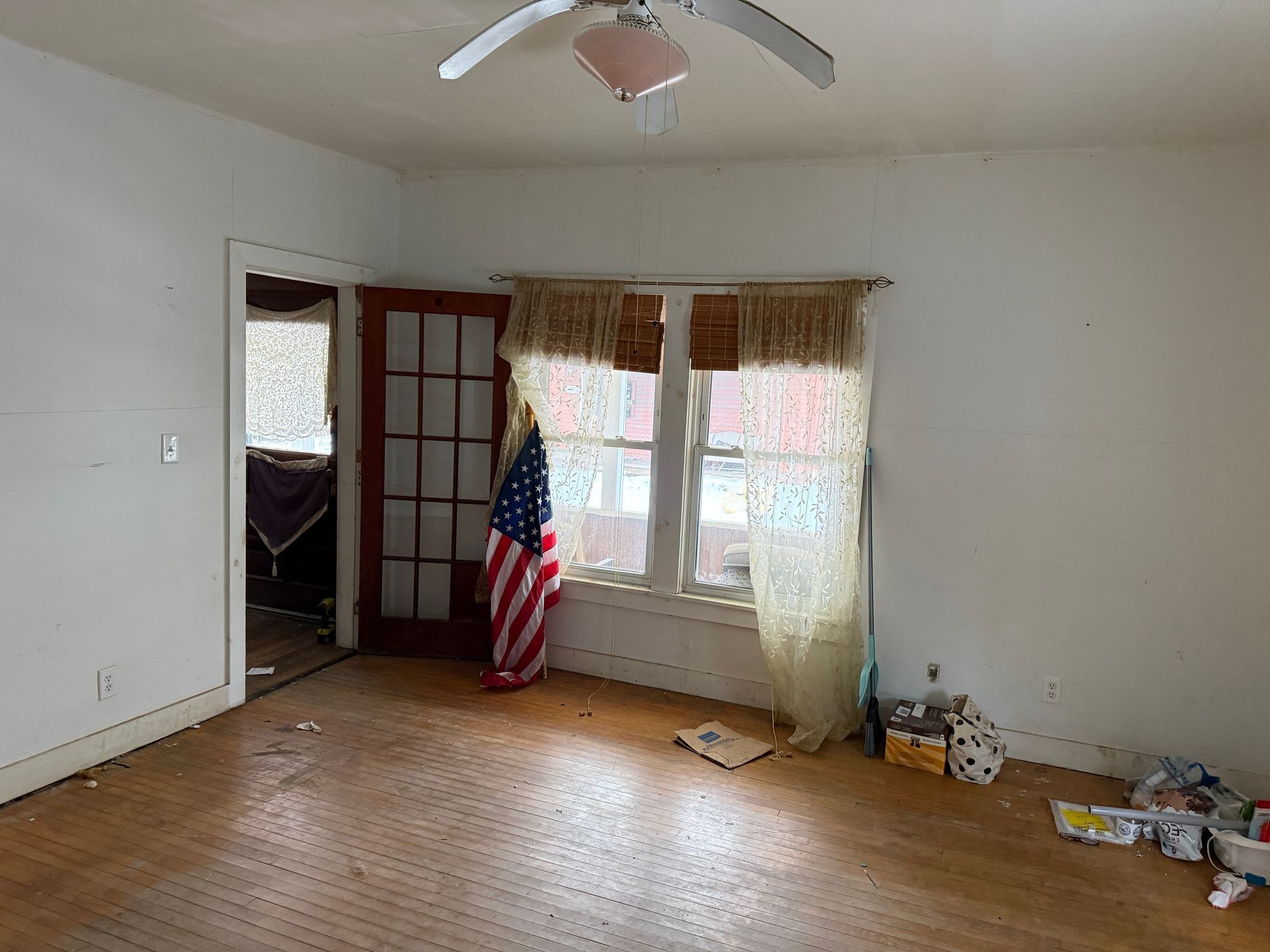 An empty room with hardwood floors, a ceiling fan, a window with sheer curtains, a doorway.