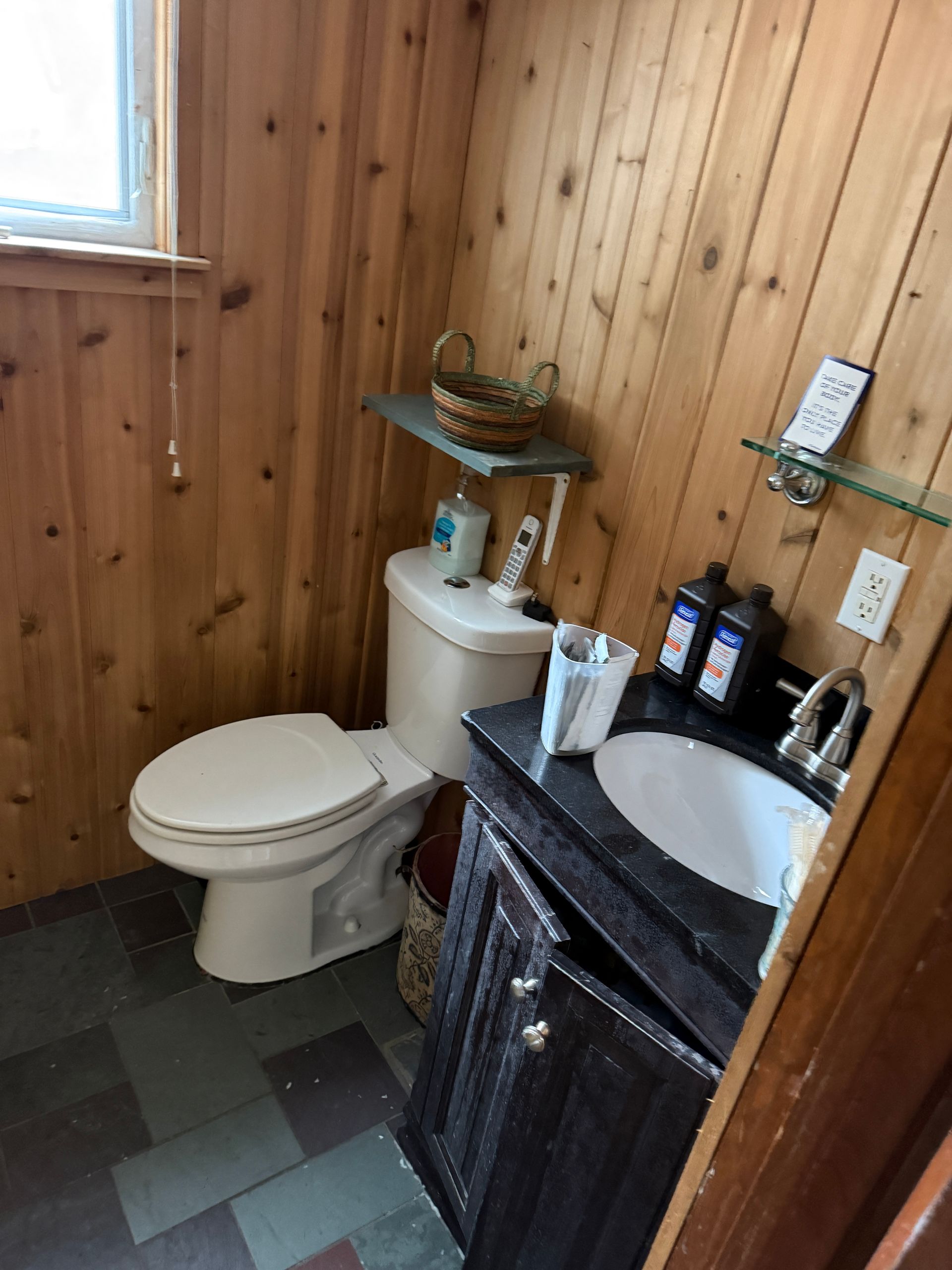A bathroom with wood-paneled walls, a white toilet, and a dark vanity with a white sink on a gray tiled floor.