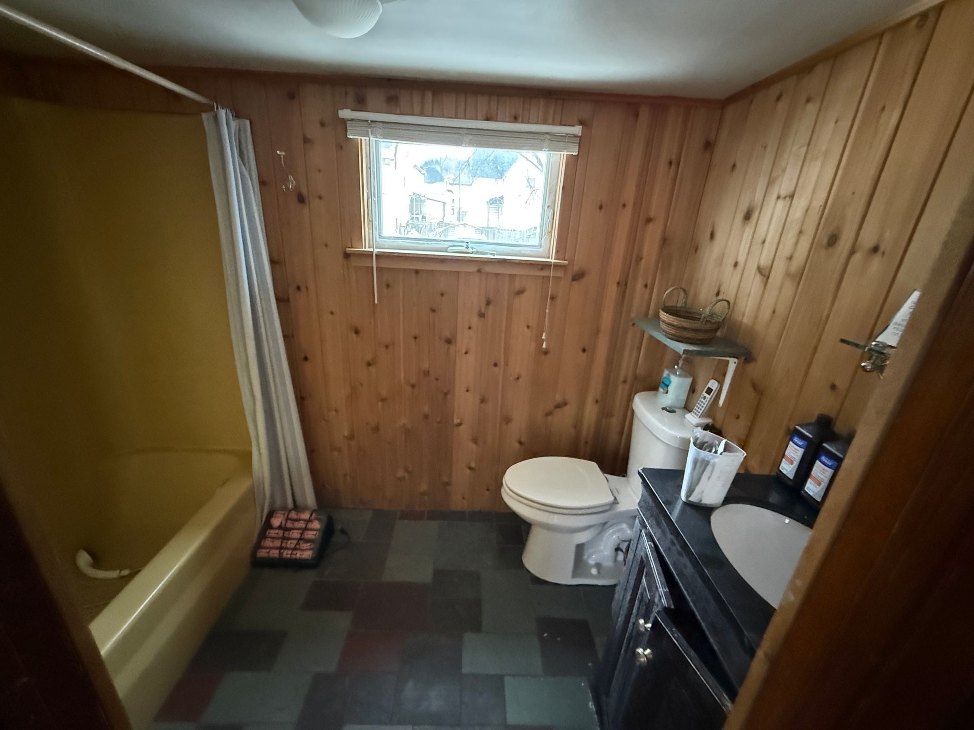 Bathroom with wood-paneled walls, yellow shower-tub, toilet, and dark vanity cabinet on a multi-toned tiled floor.